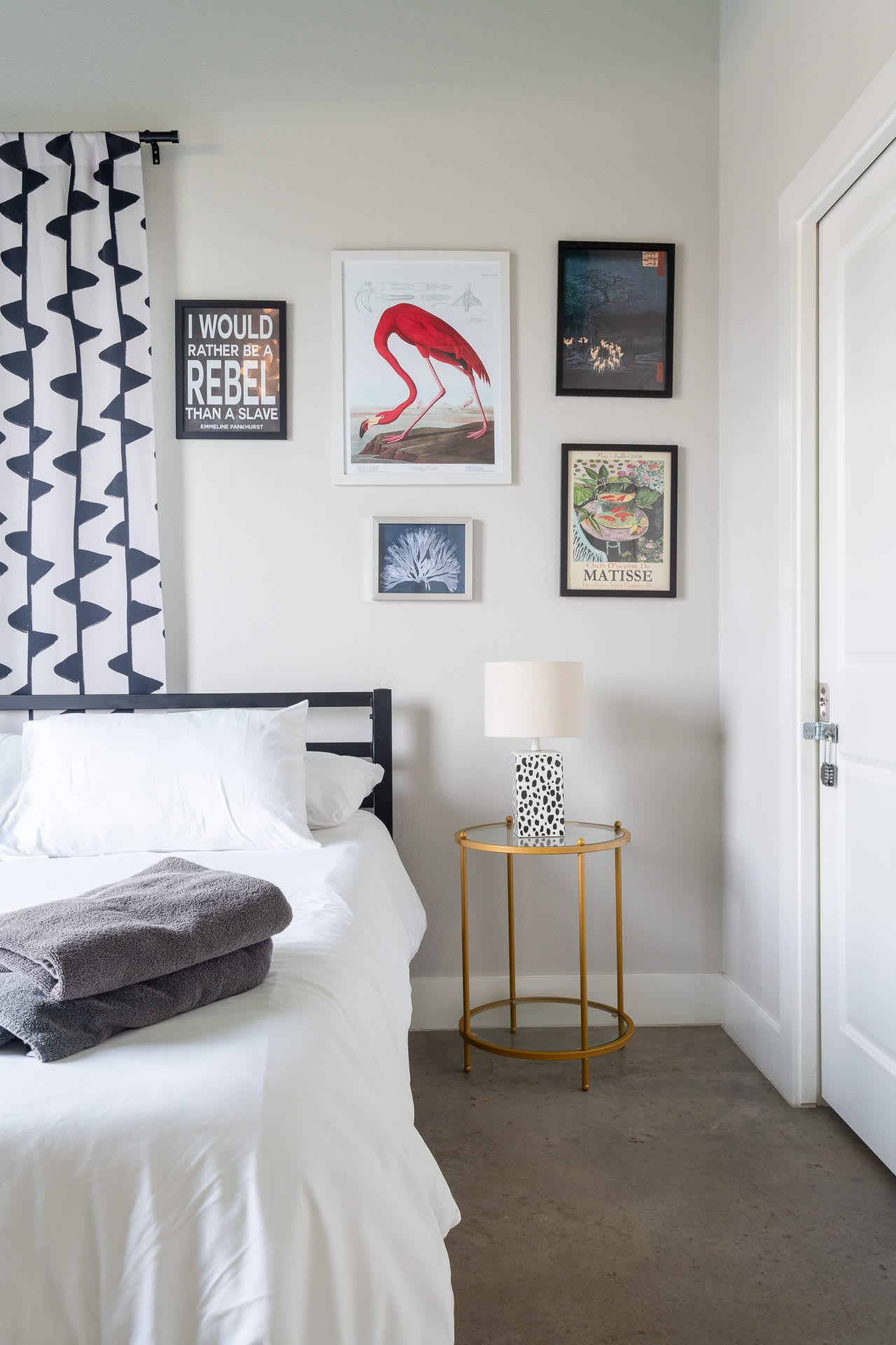 Bright bedroom featuring a black metal bed frame with white bedding, a gold-framed nightstand with a spotted lamp, and a gallery wall with diverse art prints, including a large red flamingo.