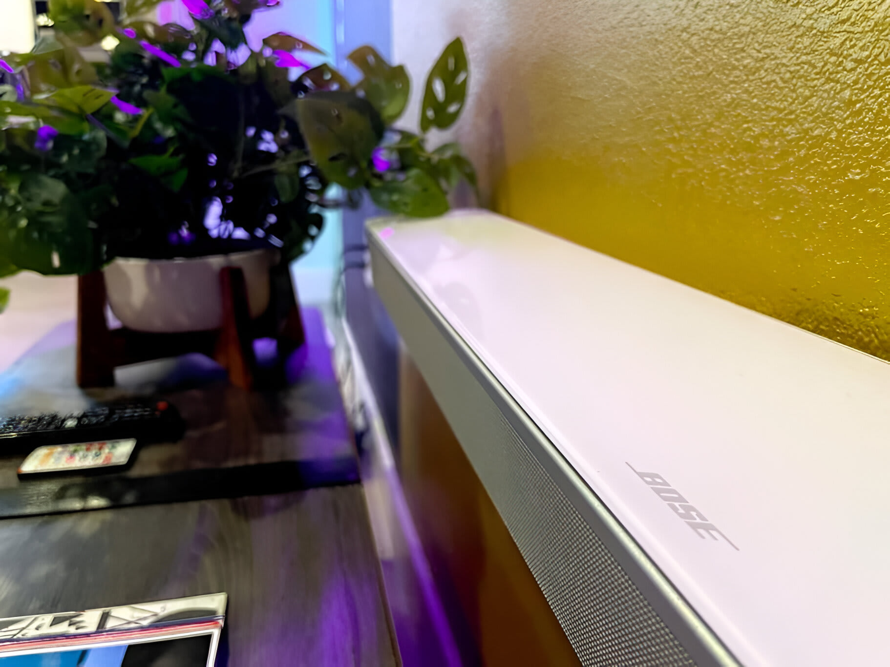A close-up view of a white Bose soundbar resting on a dark wooden media console, with a vibrant green potted plant and a yellow wall visible in the soft background.