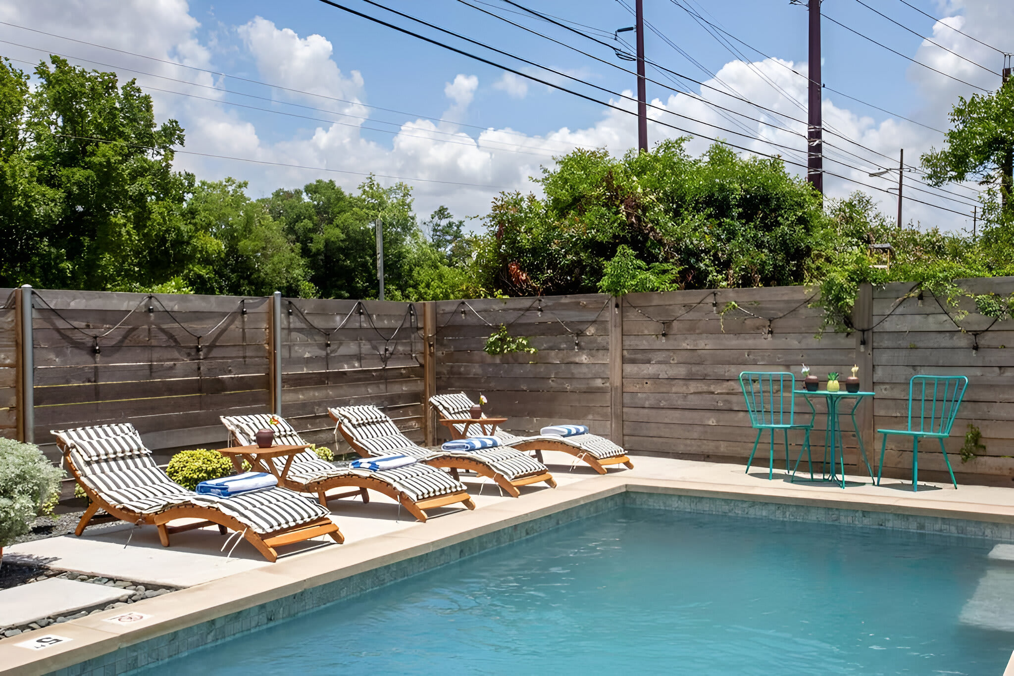 A serene outdoor swimming pool is lined with several striped lounge chairs on a paved area, surrounded by dark gravel and a tall wooden fence. A small table with two chairs is visible in the background, offering an additional seating area.