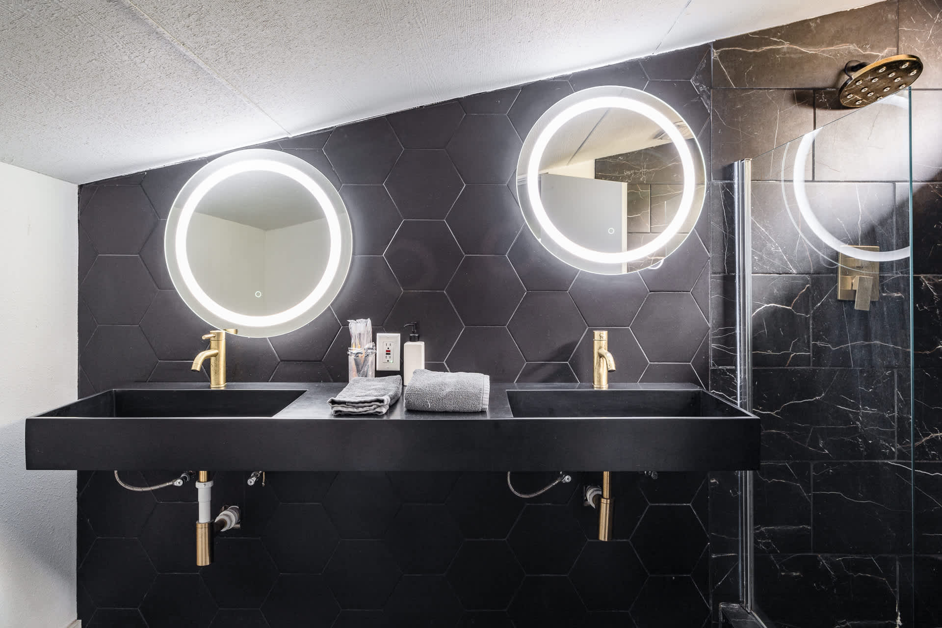 A stylish bathroom features a double black floating vanity with gold faucets and two round illuminated mirrors, set against a black hexagonal tile wall.