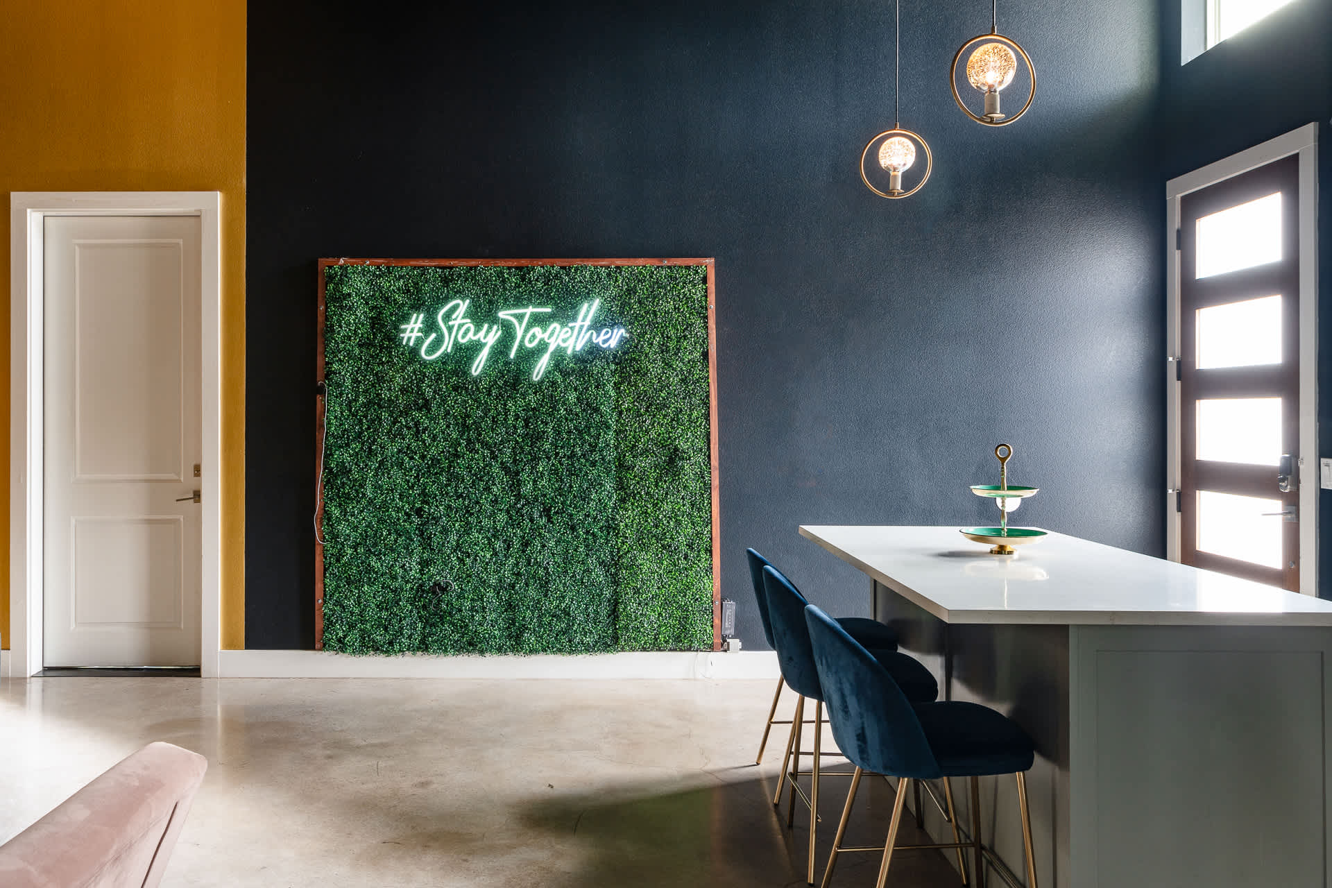 A stylish entryway or open-concept living area features a large green wall with a neon "#StayTogether" sign, adjacent to a kitchen island with two blue velvet bar stools. The space has dark blue and yellow accent walls, with modern pendant lighting.