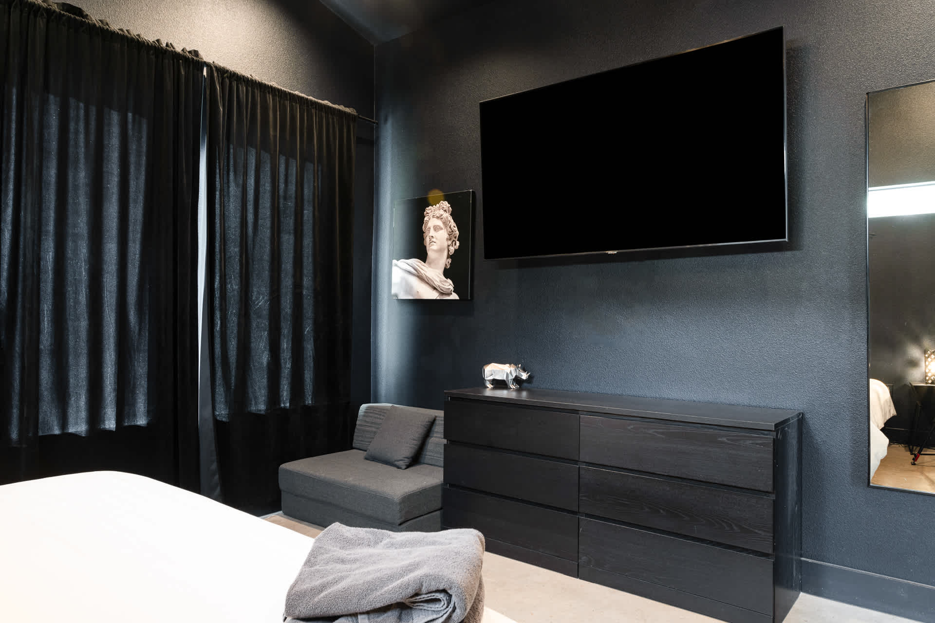 A dark-themed bedroom features a large wall-mounted flat-screen TV above a black dresser, with a grey chaise lounge chair in the corner. Dark curtains cover a window, and a framed bust artwork is visible.
