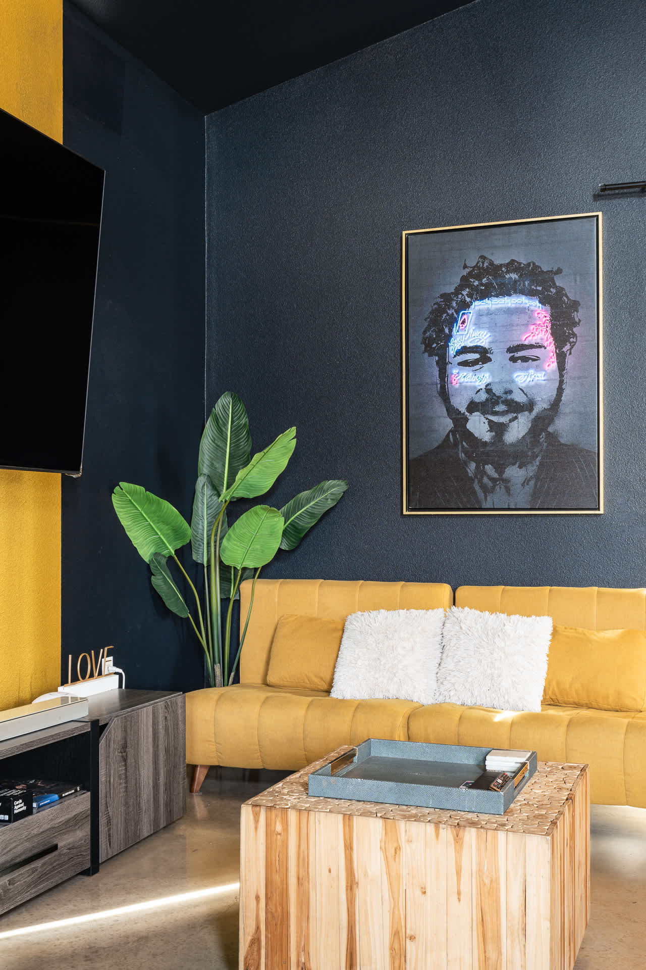 A modern living room features a vibrant yellow sofa with white throw pillows, dark blue textured walls, and a large flat-screen TV mounted above a wooden media console. A neon-lit portrait of Post Malone hangs on the wall, adding a pop of color.