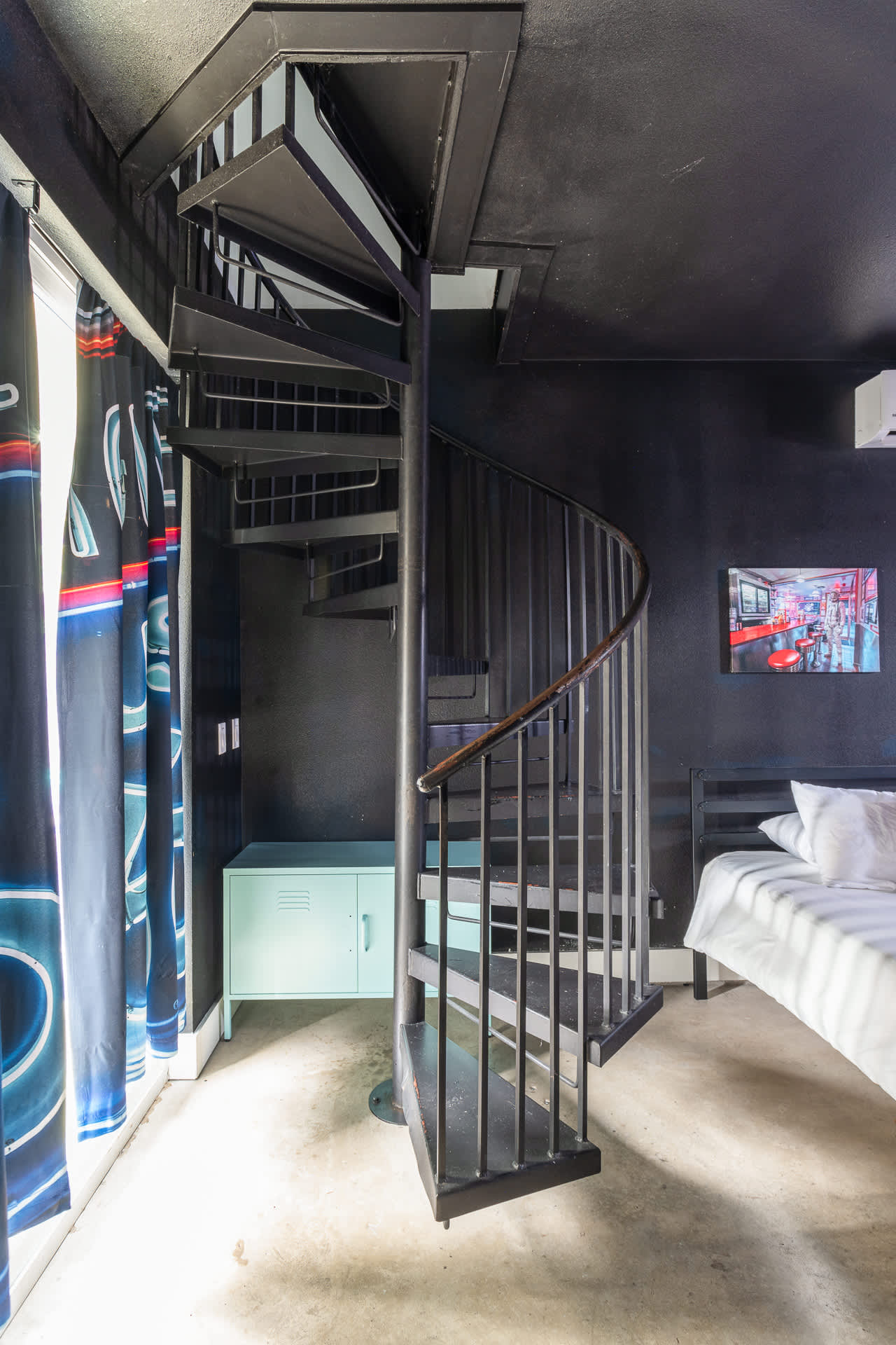 View of a black metal spiral staircase with wooden handrails, leading up from a dark-walled bedroom with concrete floors and a light blue cabinet visible near the window.