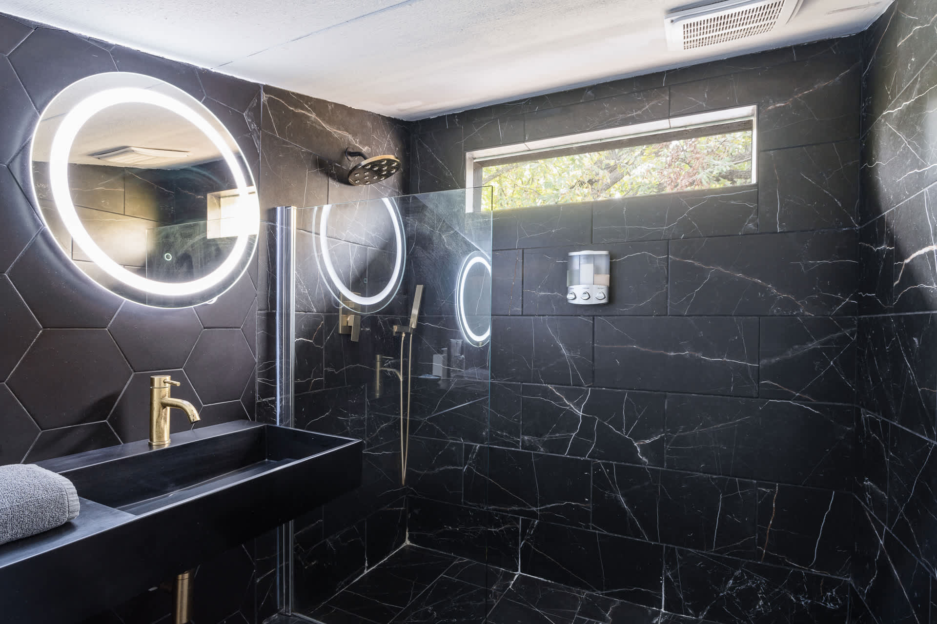 A modern bathroom with a walk-in shower featuring black marble-patterned tiles and a glass partition. A black floating vanity with a gold faucet and an illuminated mirror is visible on the left.