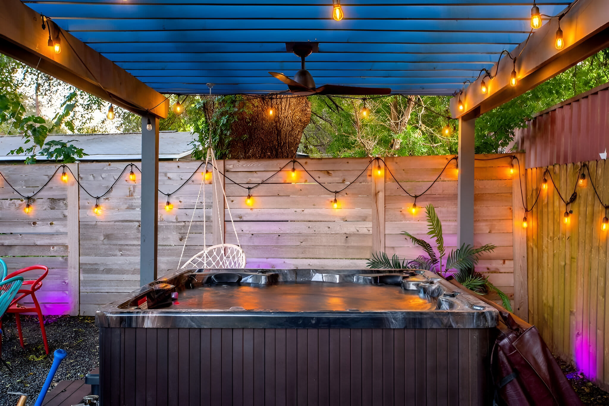 An evening view of an outdoor hot tub under a blue pergola, illuminated by warm string lights and purple accent lighting. A white hanging chair is visible next to the hot tub, creating a cozy and inviting atmosphere.