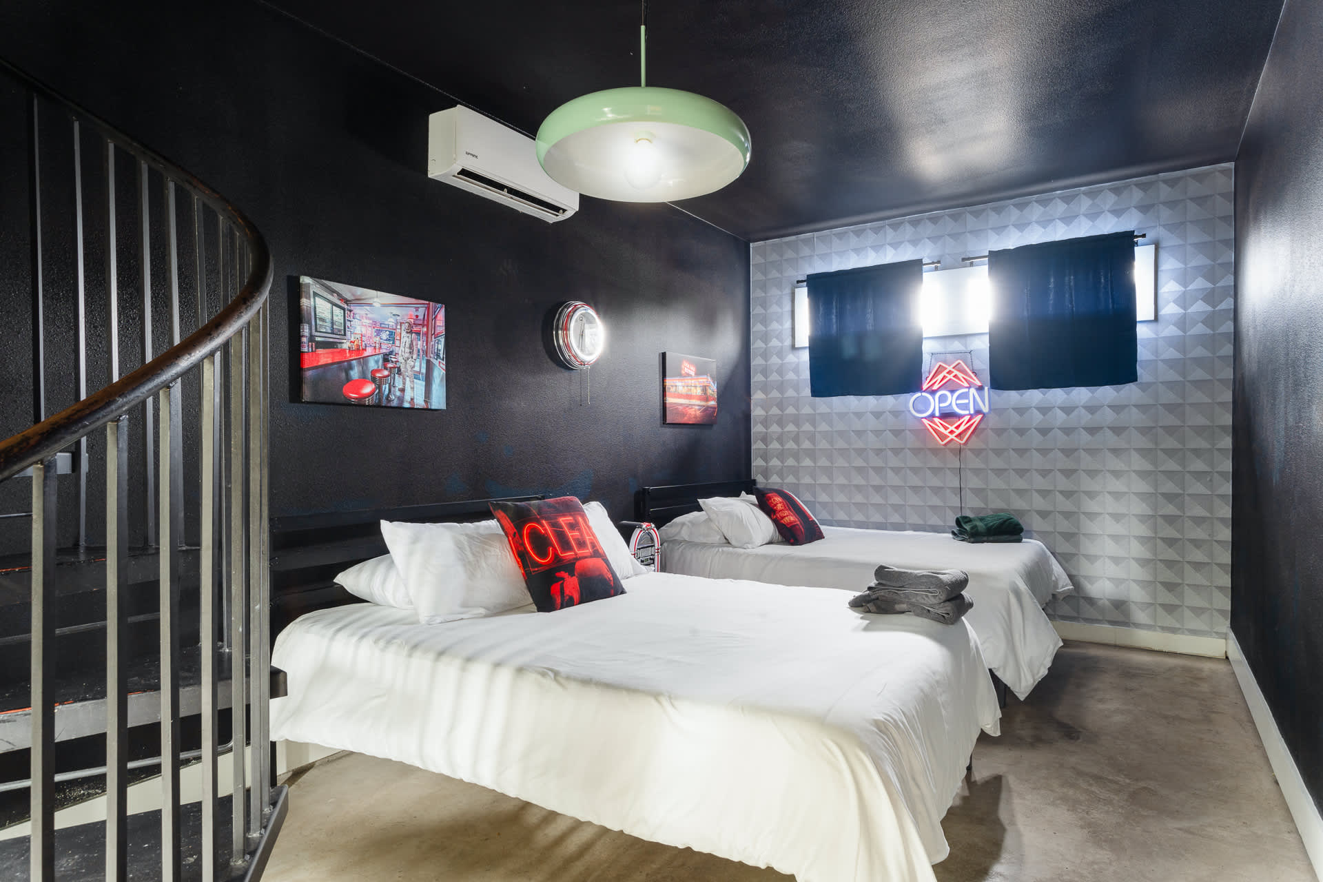 Dark-themed bedroom with two single beds featuring white bedding, black walls, a geometric patterned accent wall, and a vibrant 'OPEN' neon sign, with a spiral staircase visible on the left.