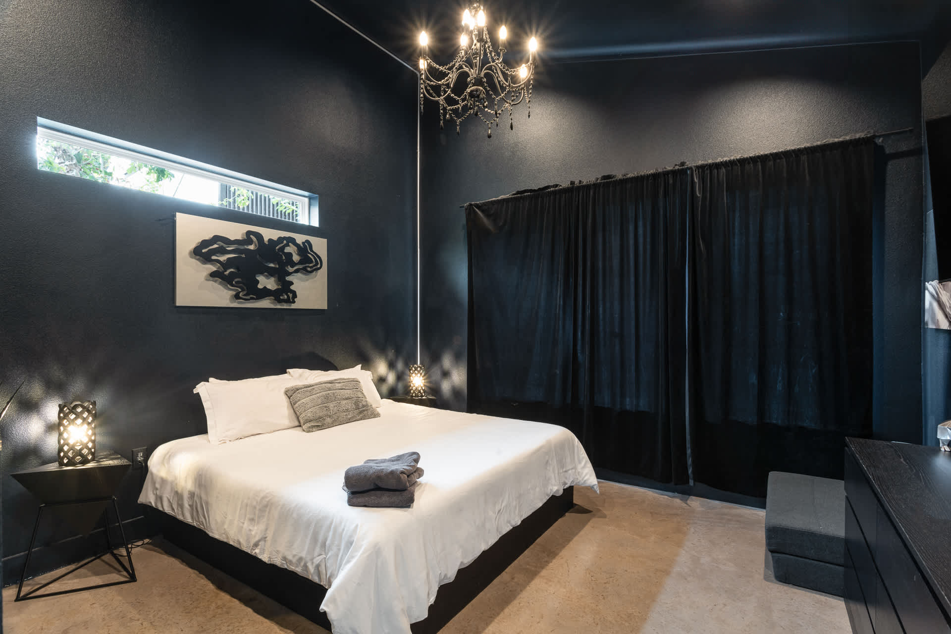 A moody bedroom features a queen-sized bed with white linens, dark textured walls, and a sparkling crystal chandelier hanging from the ceiling. A long horizontal window is positioned above a modern abstract artwork.