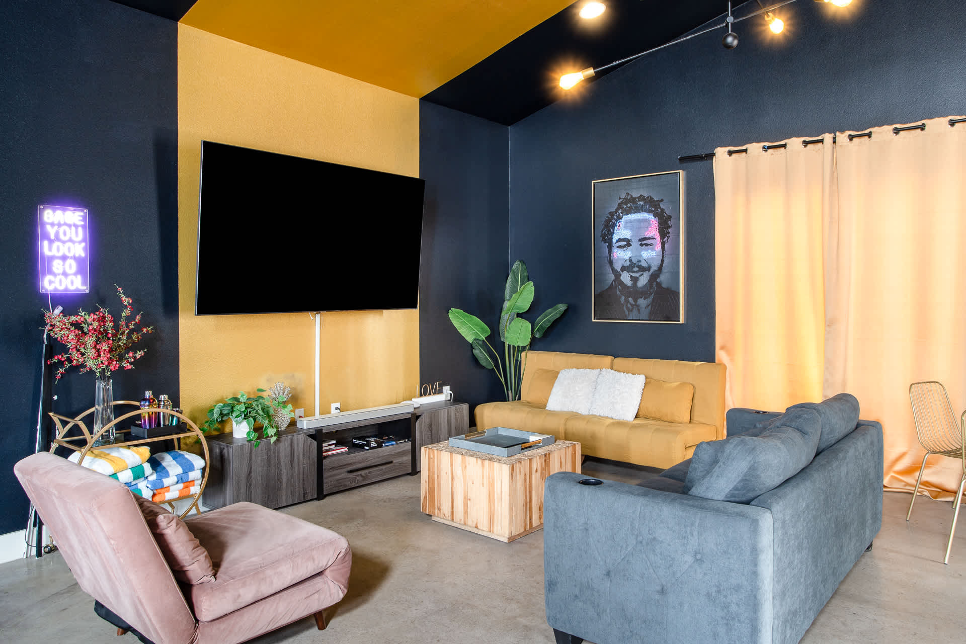 A vibrant living room features a large wall-mounted TV against a two-tone yellow and dark blue wall, with a yellow sofa, a gray sofa, and a light pink armchair arranged around a wooden coffee table. A neon sign and a large Post Malone portrait add to the eclectic decor.