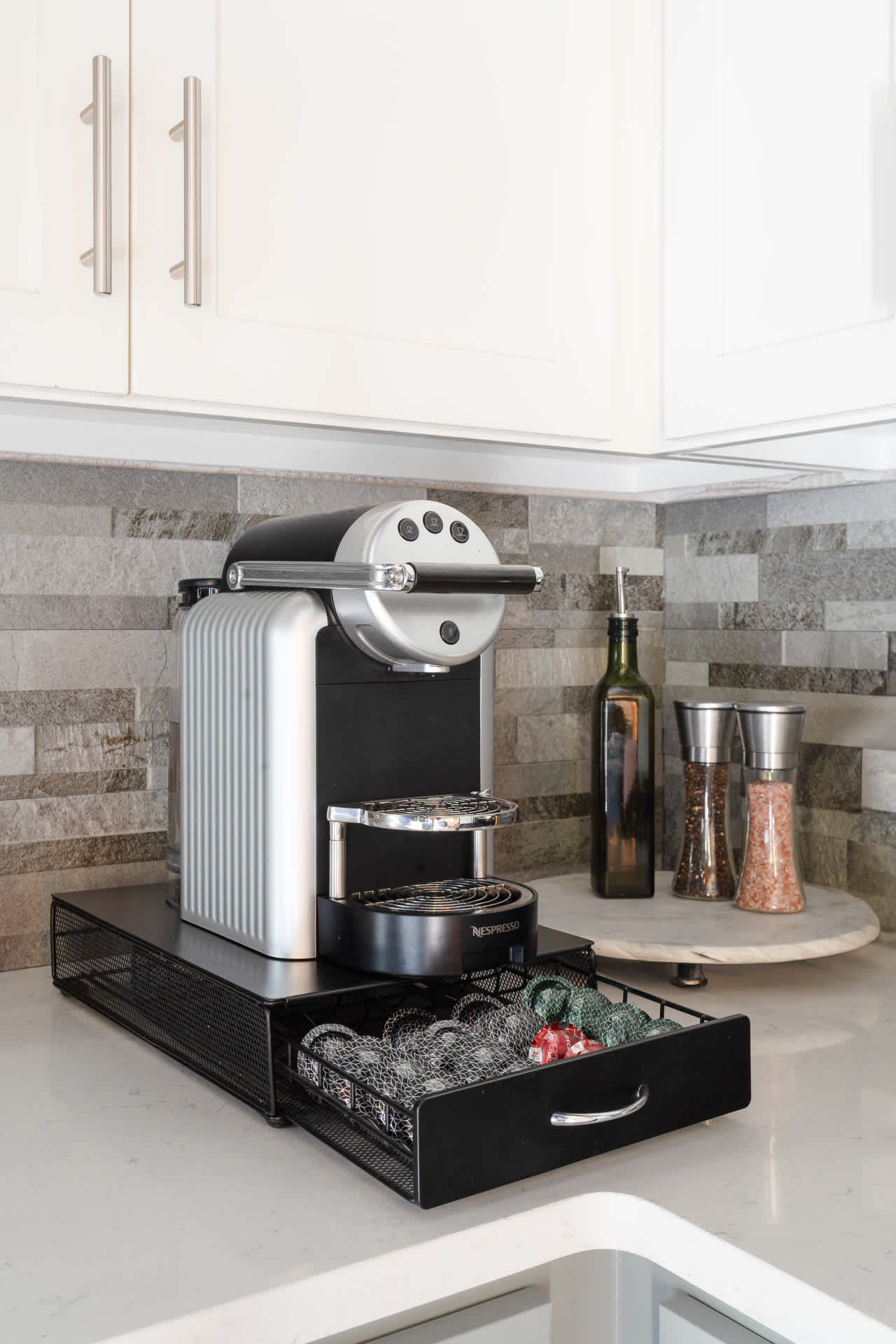 A sleek Nespresso coffee machine sits on a white quartz countertop with a black mesh drawer organizer, set against a grey subway tile backsplash. White shaker cabinets are visible above.