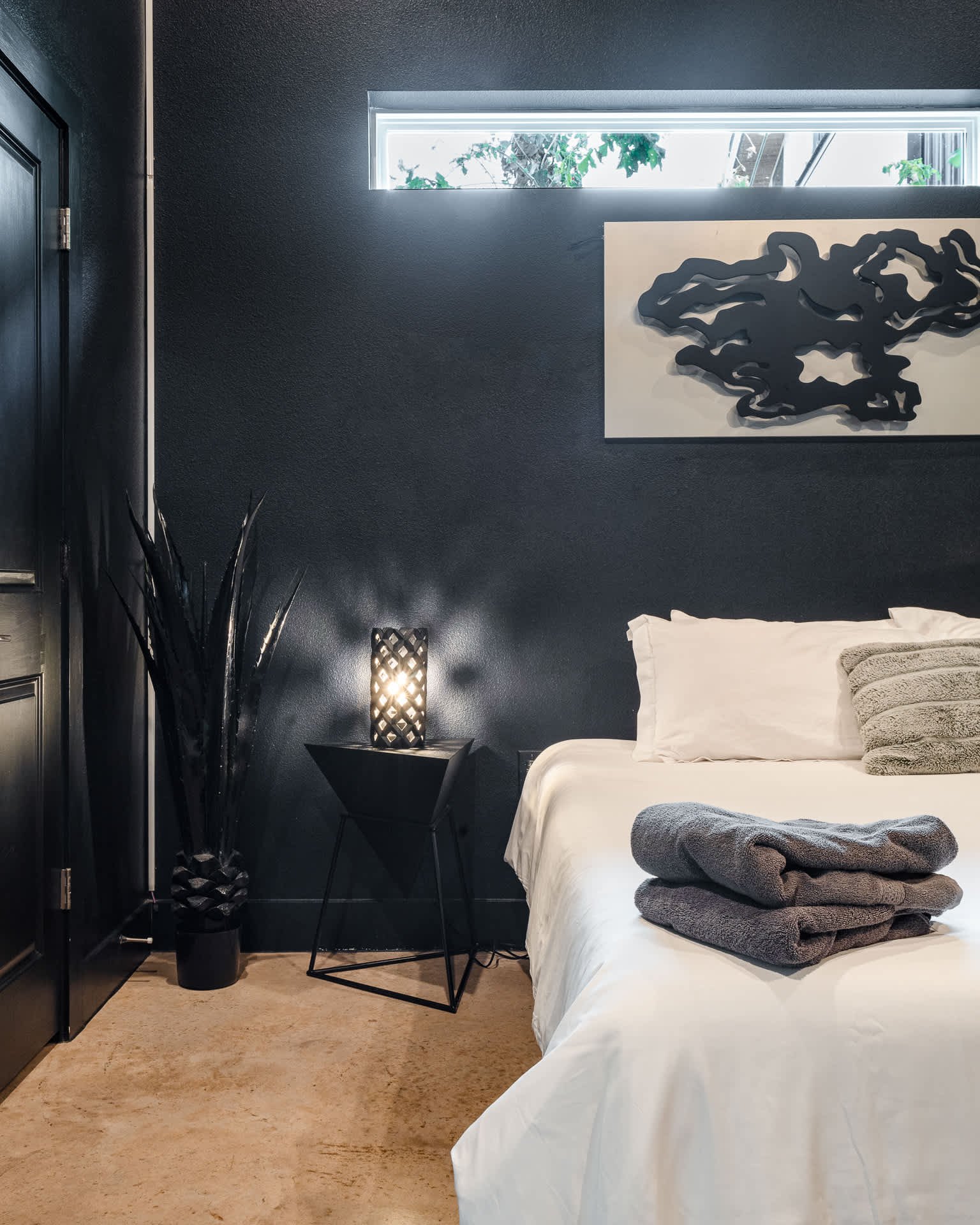 A close-up of a modern black nightstand with a geometric patterned lamp next to a bed with white linens and folded grey towels. A tall dark plant in a textured pot stands by the door.