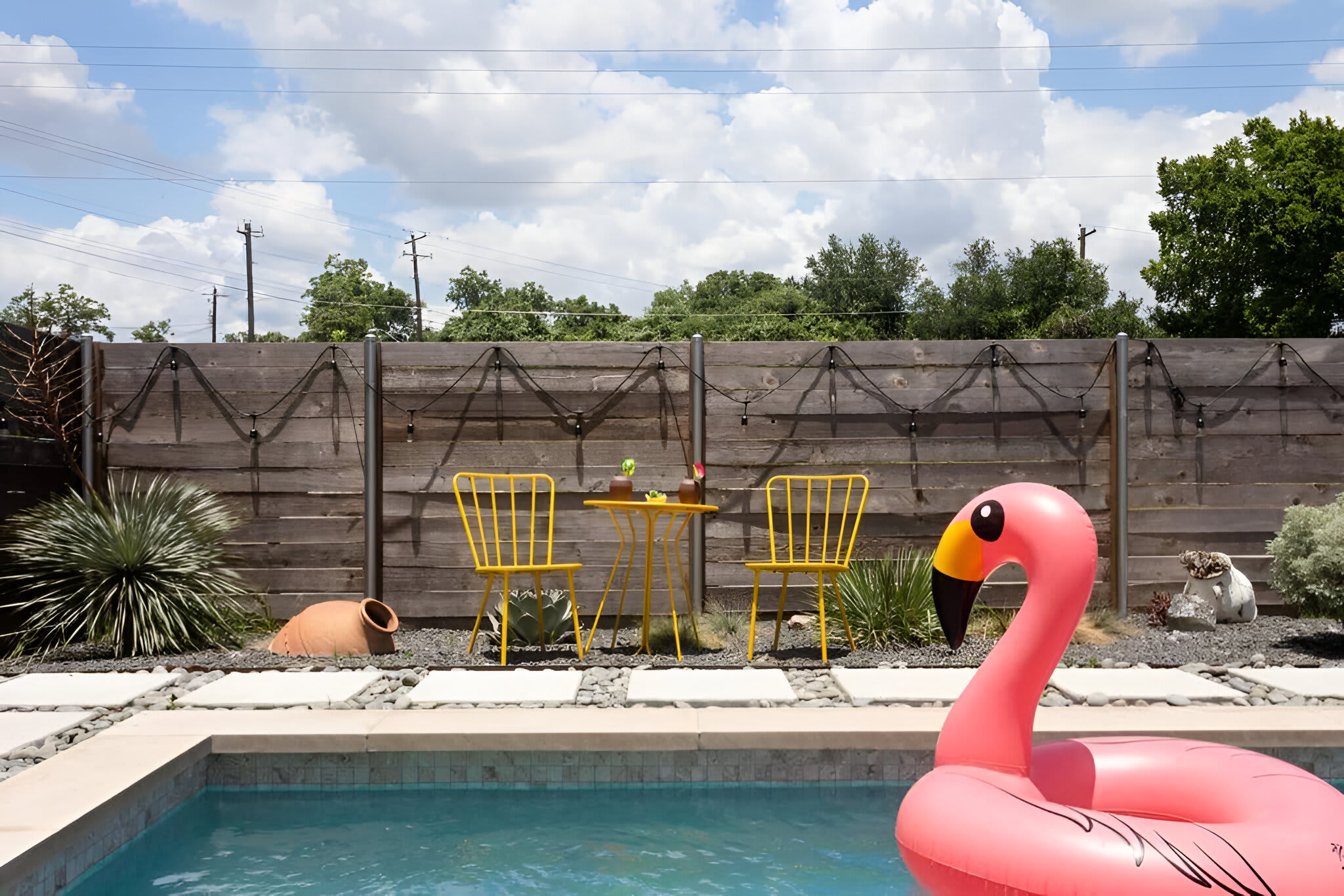 A close-up view of an outdoor swimming pool shows a large pink flamingo float in the foreground, with a small yellow table and two matching chairs set on a gravel and paver patio. A wooden fence and lush greenery frame the background under a bright sky.