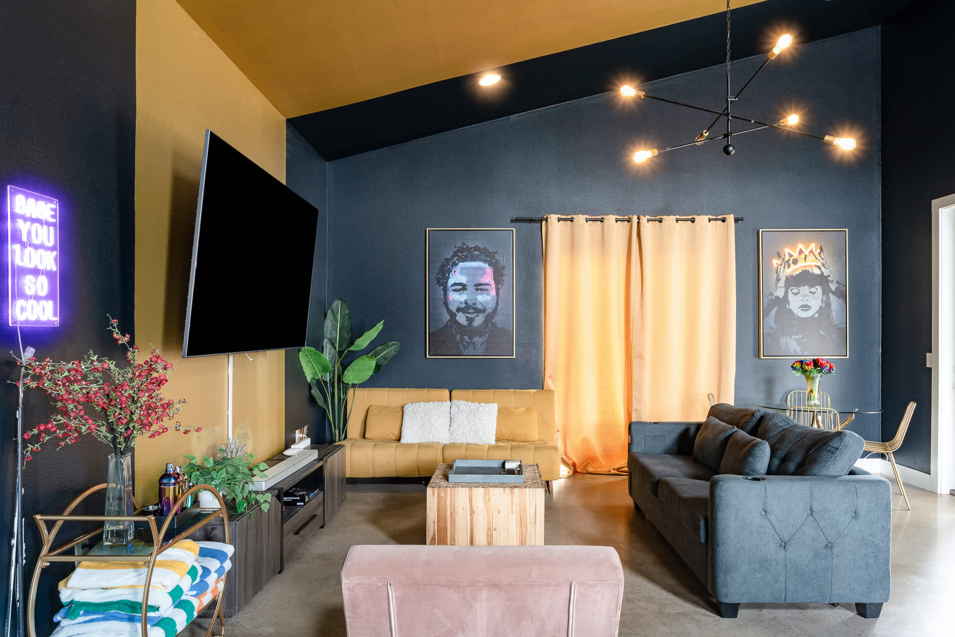 A spacious living room with a high vaulted ceiling features a large wall-mounted TV against a yellow and dark blue accent wall, complemented by a yellow sofa, a gray sofa, and a pink armchair. A Post Malone portrait and a crown-wearing figure portrait adorn the dark blue wall.
