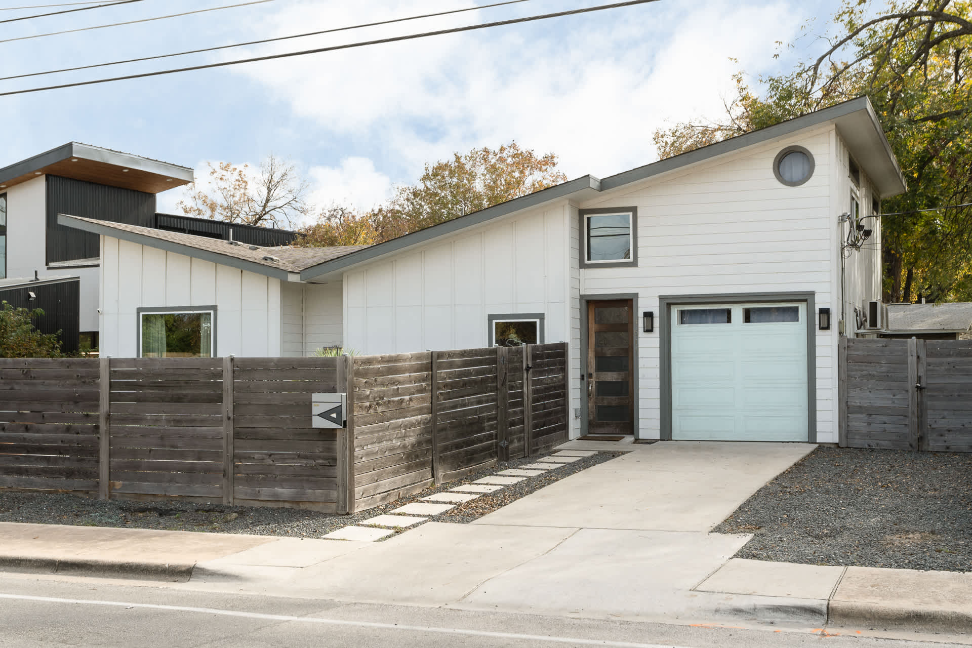 Modern white house exterior with a light blue garage door, a dark wooden fence, and a concrete driveway with stepping stones, under a partly cloudy sky.