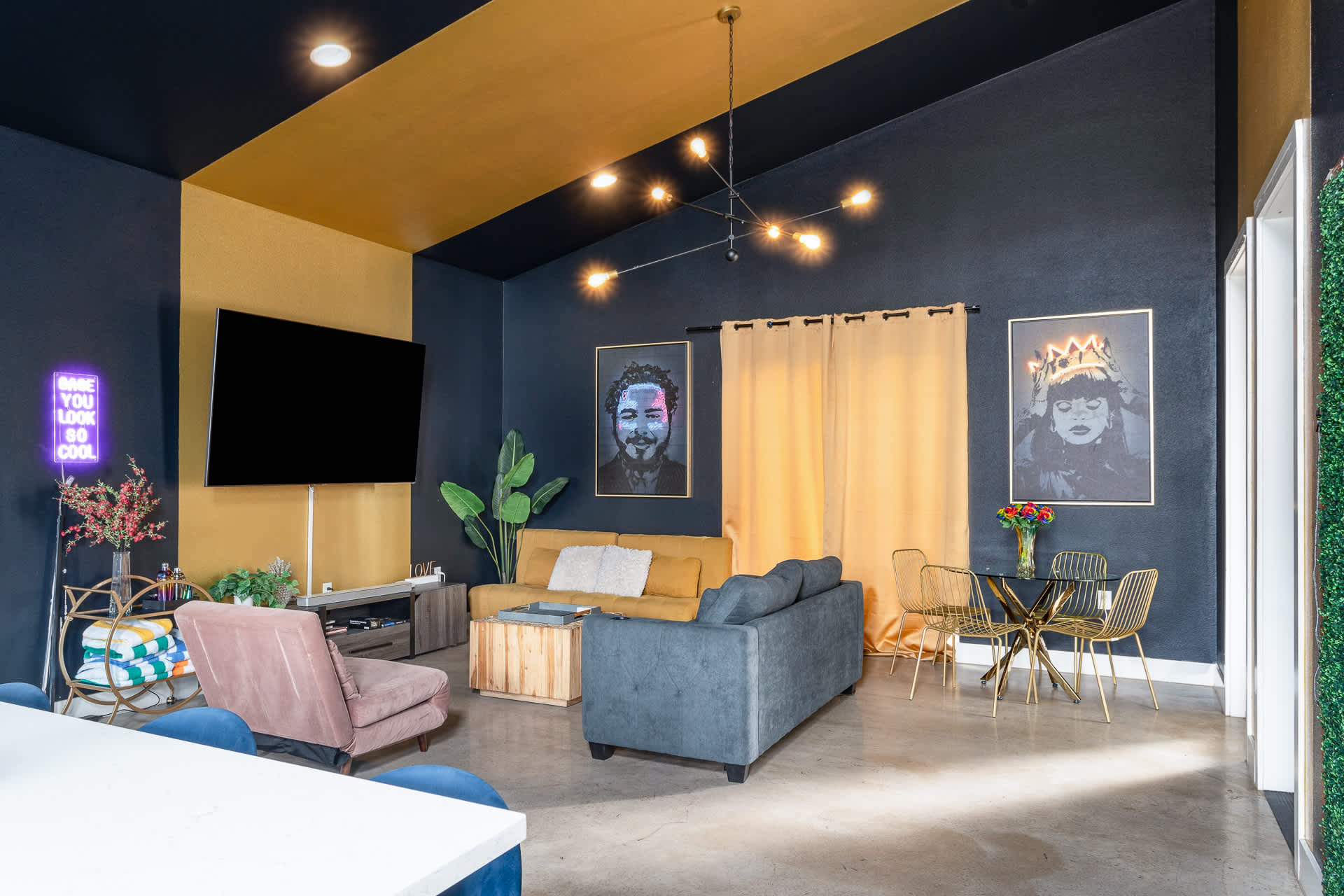 A stylish living area with a striking black and yellow striped ceiling, featuring a yellow sofa, a grey loveseat, and a small dining set with gold chairs. A large TV, neon signs, and framed art pieces adorn the dark walls.