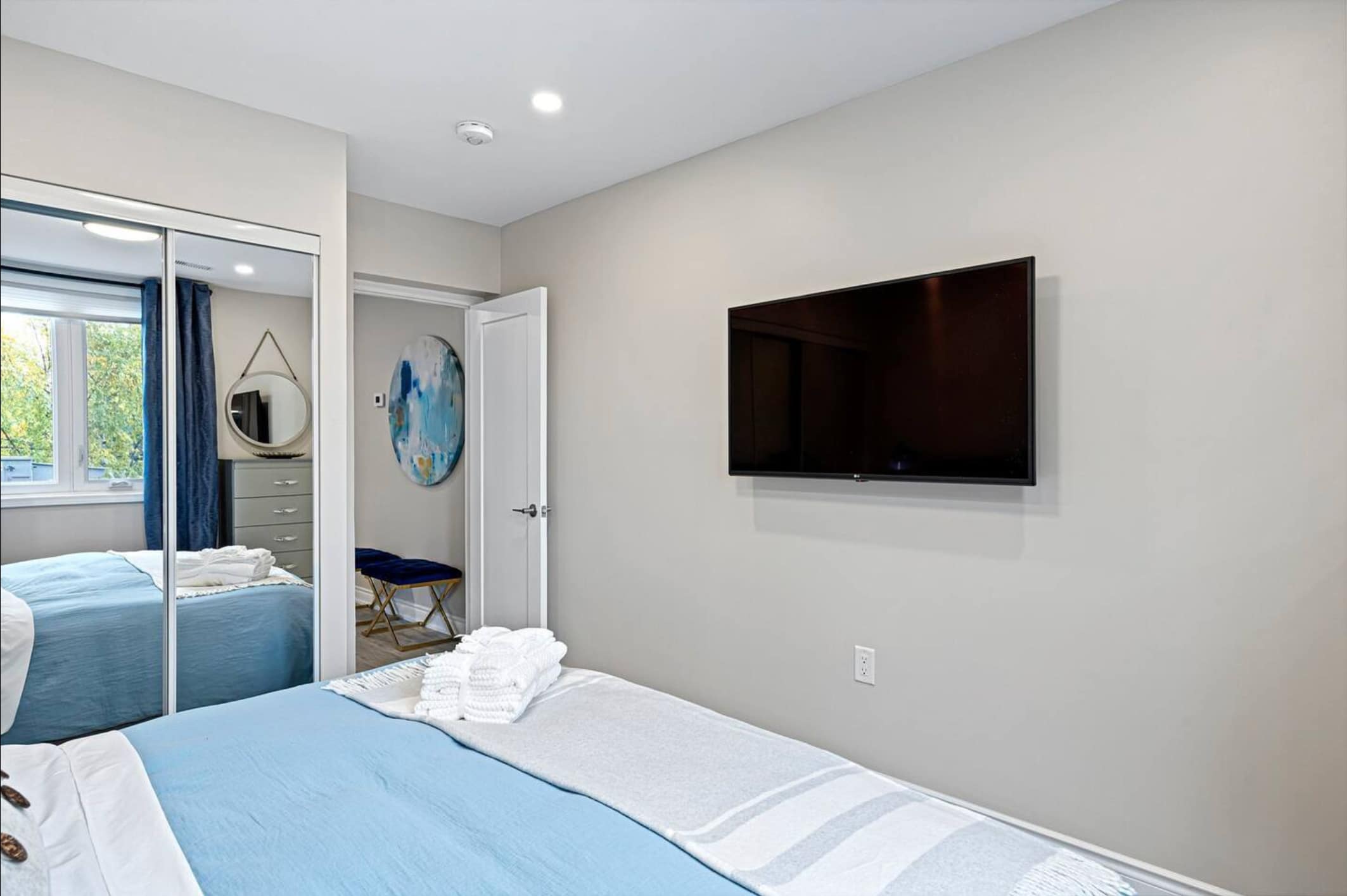 A bedroom with light gray walls features a wall-mounted flat-screen TV, a bed with blue and gray bedding, and a mirrored closet reflecting a window with blue curtains. A doorway leads to another room with a blue accent chair.