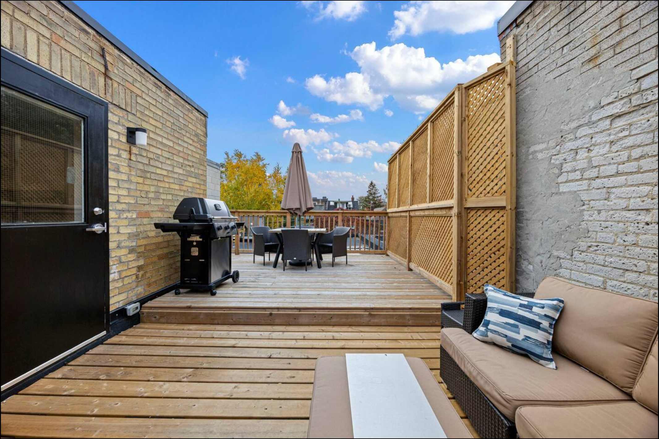 A spacious wooden deck featuring a BBQ grill, an outdoor dining set with an umbrella, and a lounge seating area with cushions. A privacy lattice screen and a brick wall are also visible under a blue sky.