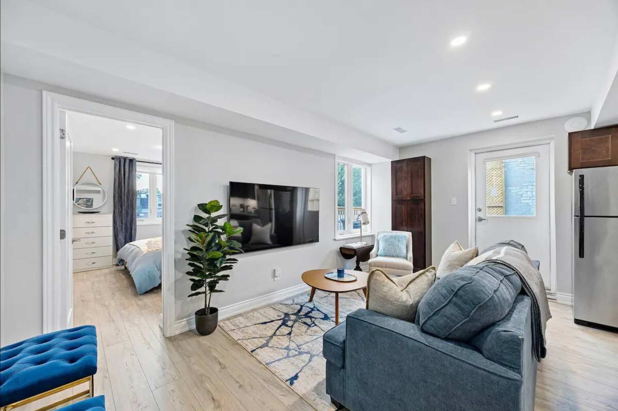 A bright living room with a large wall-mounted TV, a blue sofa, and a round wooden coffee table. A doorway on the left leads to a bedroom, and a kitchen area is visible to the right.