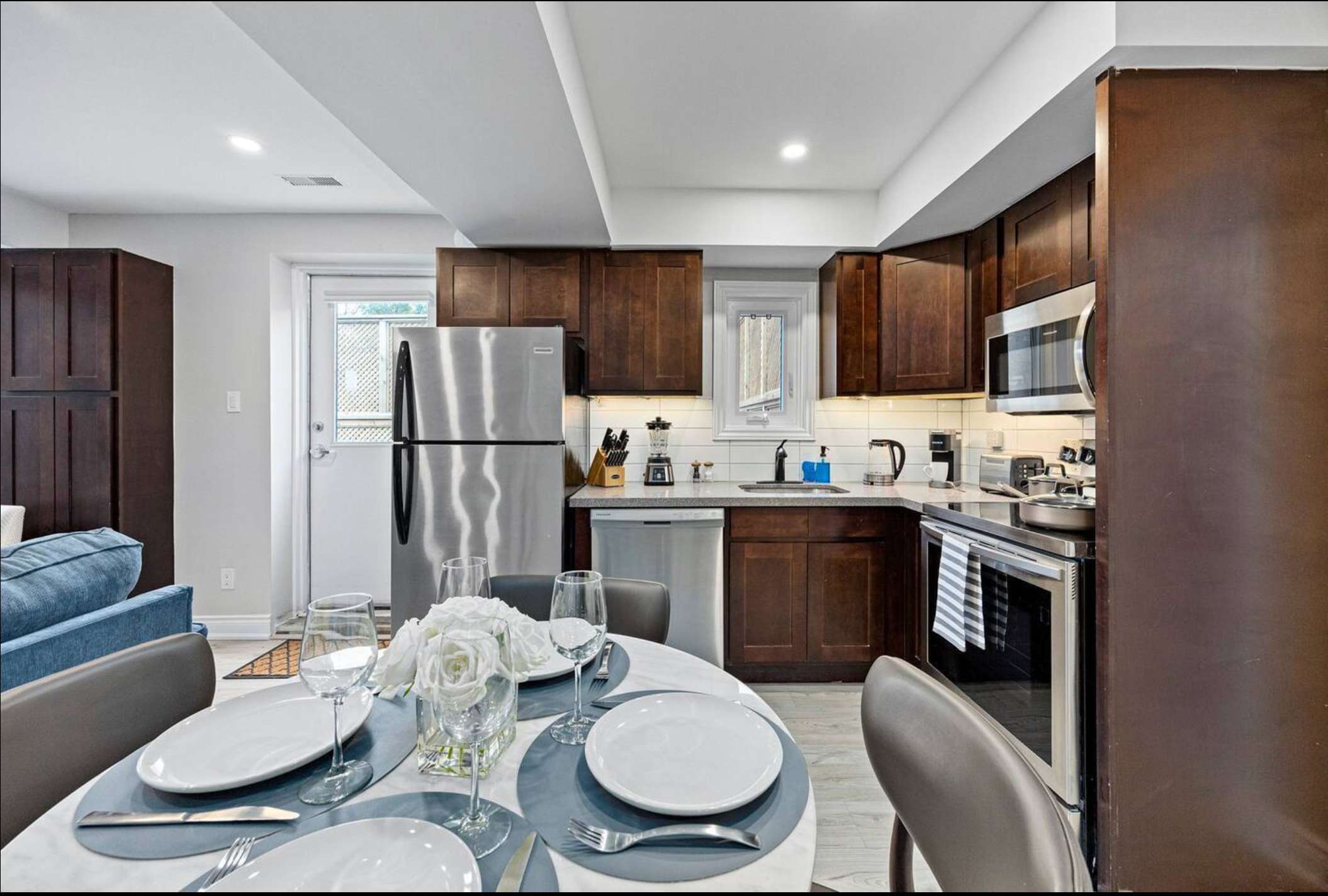 An open-concept kitchen and dining area features dark wood cabinets, stainless steel appliances including a refrigerator, dishwasher, and microwave, and a round dining table set for four. White subway tiles line the backsplash, and a door leads to an outdoor area.