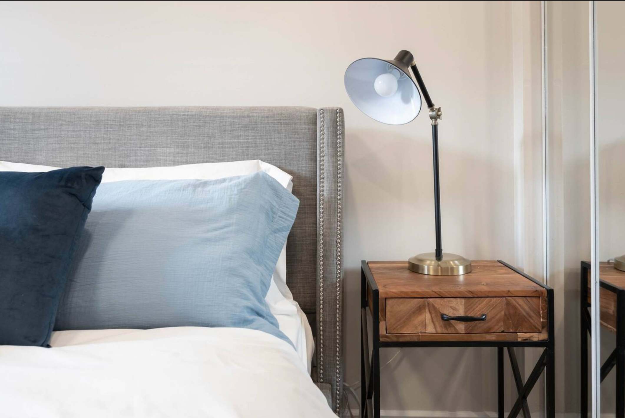 A cozy bedroom features a gray upholstered headboard with nailhead trim, light blue and dark blue pillows, and a wooden nightstand with a black and brass desk lamp. A mirrored closet door is visible on the right.