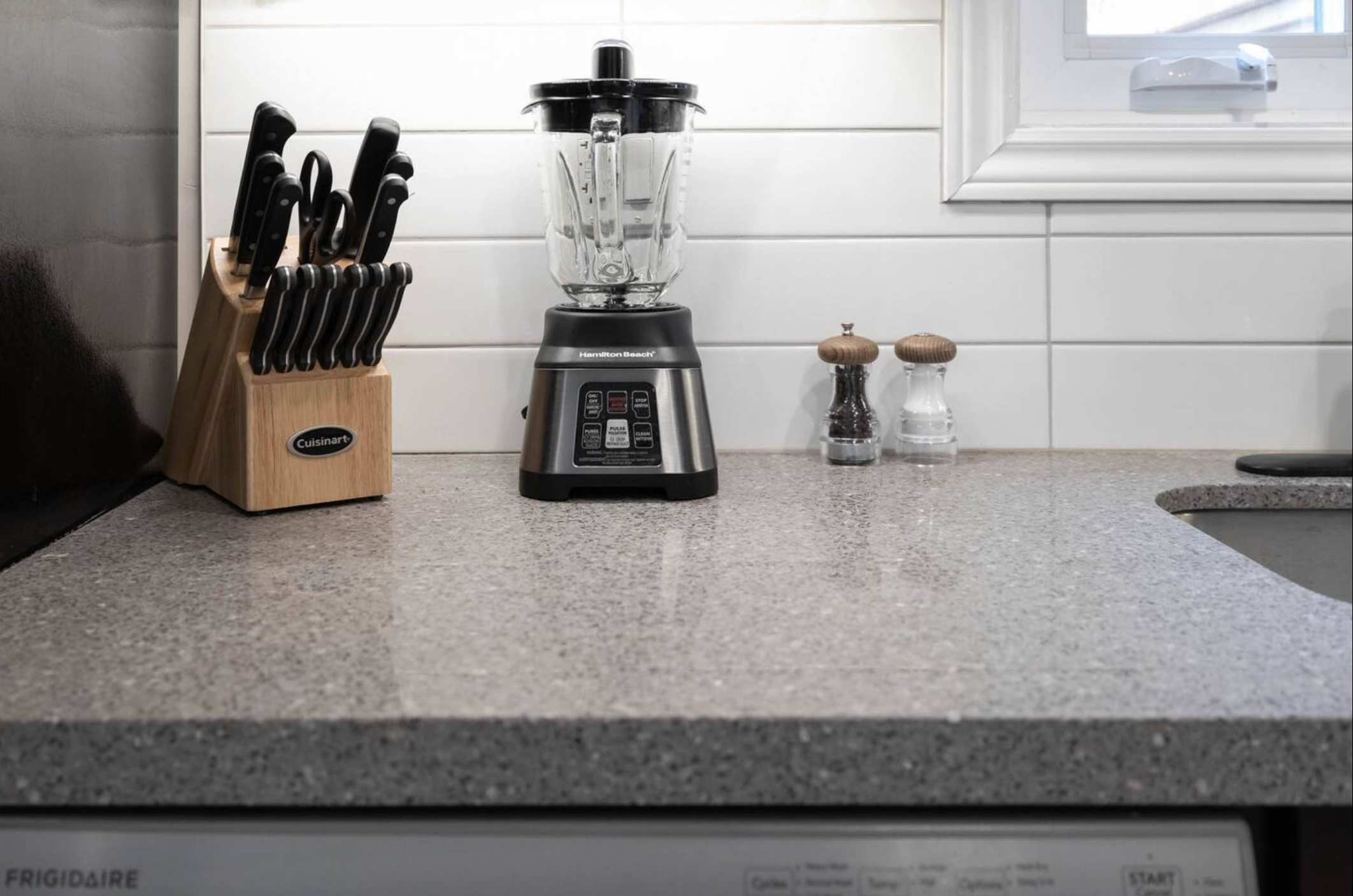 A kitchen countertop displays a wooden knife block, a Hamilton Beach blender, and salt and pepper grinders against a white subway tile backsplash. A window with white trim is visible in the background.