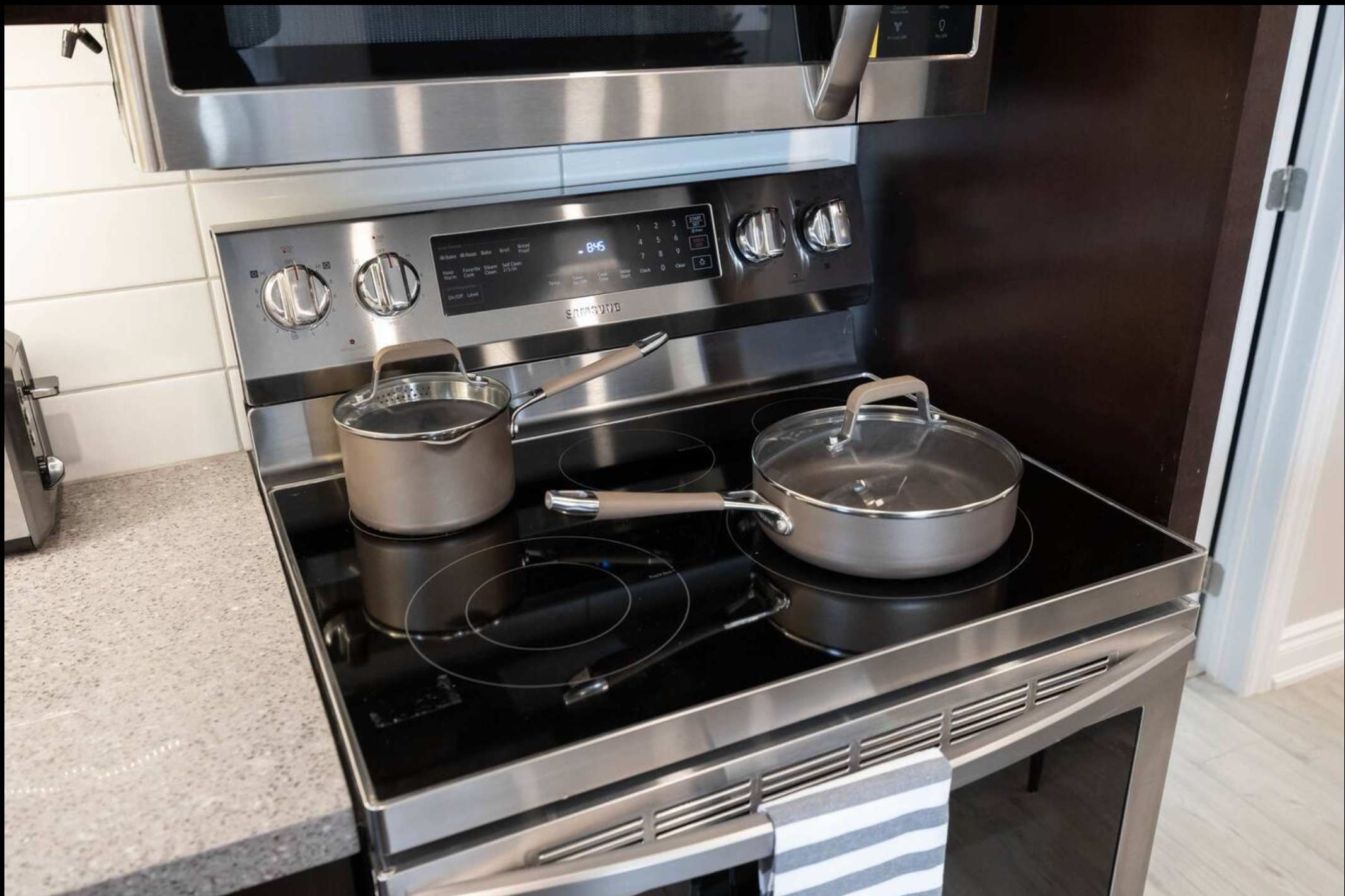 A close-up view of a modern stainless steel electric range with a black ceramic cooktop and two pots, set against a white subway tile backsplash. A microwave is visible above the stove.