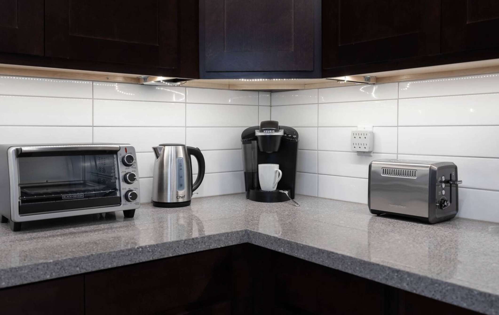 A modern kitchen corner features white subway tile backsplash, dark wood cabinets, and a speckled gray countertop with a toaster oven, kettle, coffee maker, and toaster. Under-cabinet lighting illuminates the workspace.