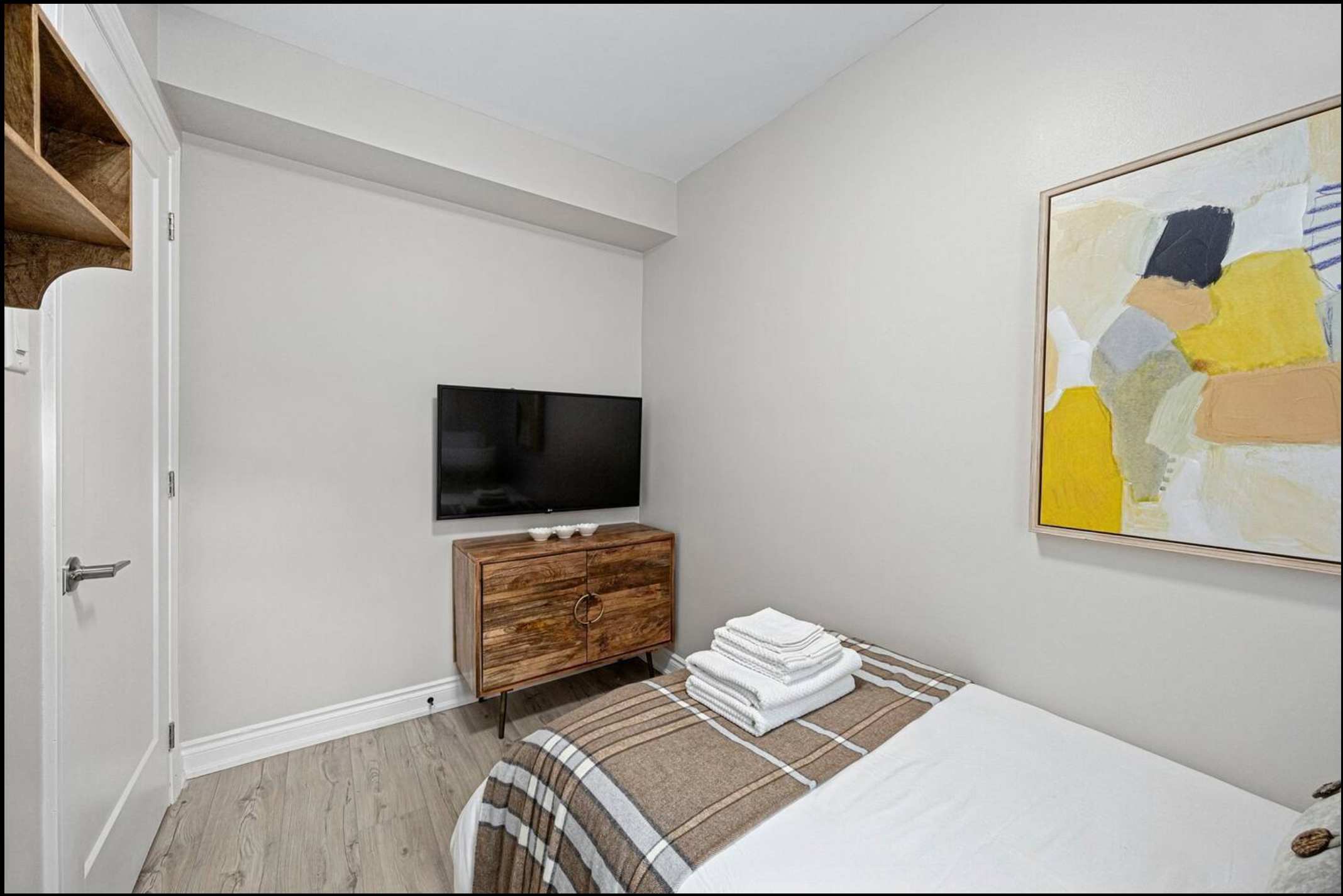 This bedroom features a wall-mounted flat-screen TV above a rustic wooden cabinet, a single bed with a brown plaid throw, and abstract artwork on the light gray wall. Light wood flooring completes the space.