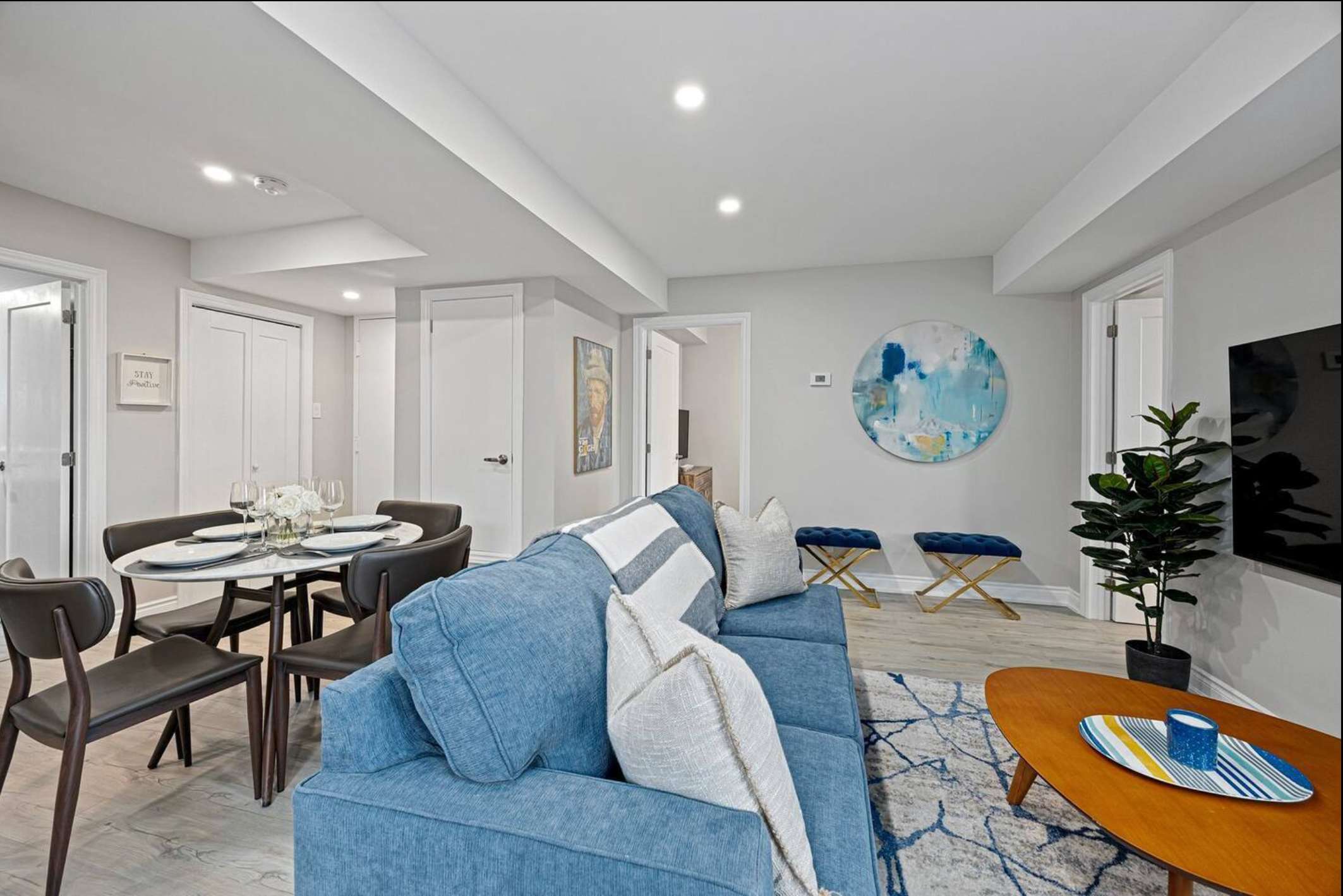 An open-concept living and dining area with a blue sofa, a round wooden coffee table, and a circular dining table set for four. Light wood floors and recessed lighting create a bright, inviting atmosphere.