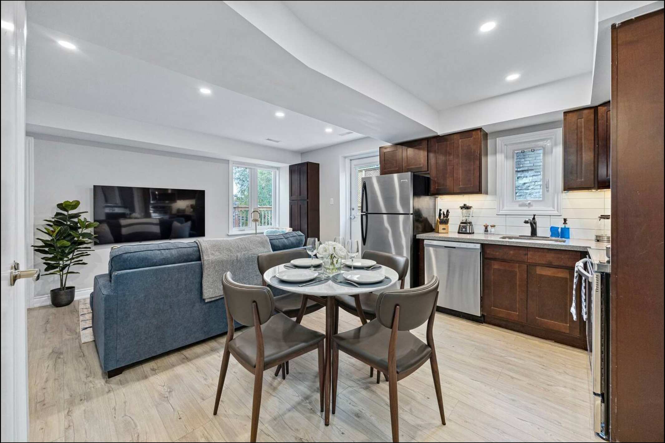 A modern open-concept kitchen with dark wood cabinets and stainless steel appliances, flowing into a dining area with a round table and chairs, and a living room with a blue sofa in the background.