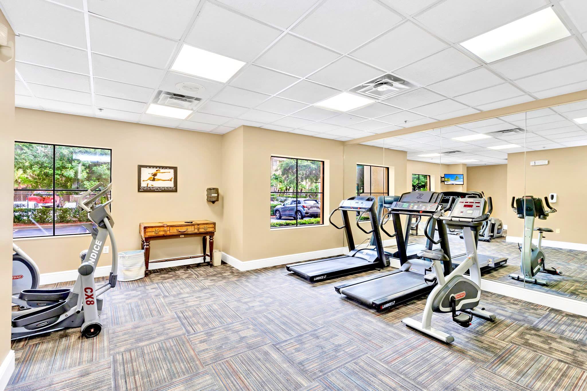 Well-lit indoor fitness center with patterned carpet, featuring two treadmills, an elliptical machine, and an exercise bike, with large windows overlooking outdoor greenery and a mirrored wall.