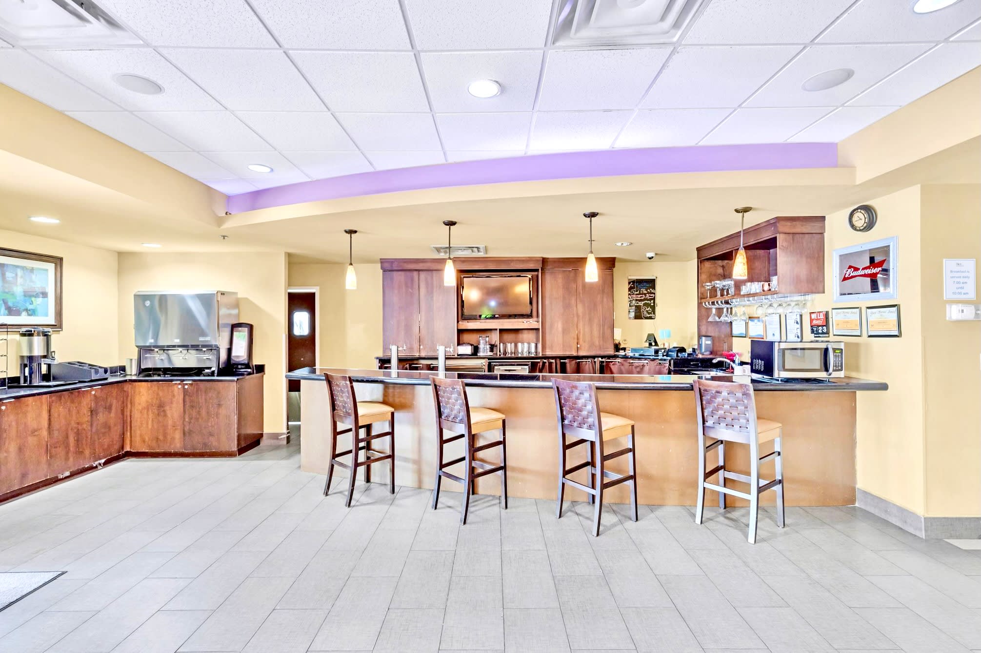 Modern bar area with a long counter and high-back bar stools, featuring wooden cabinetry, a beverage dispenser, and a television screen, under recessed lighting.