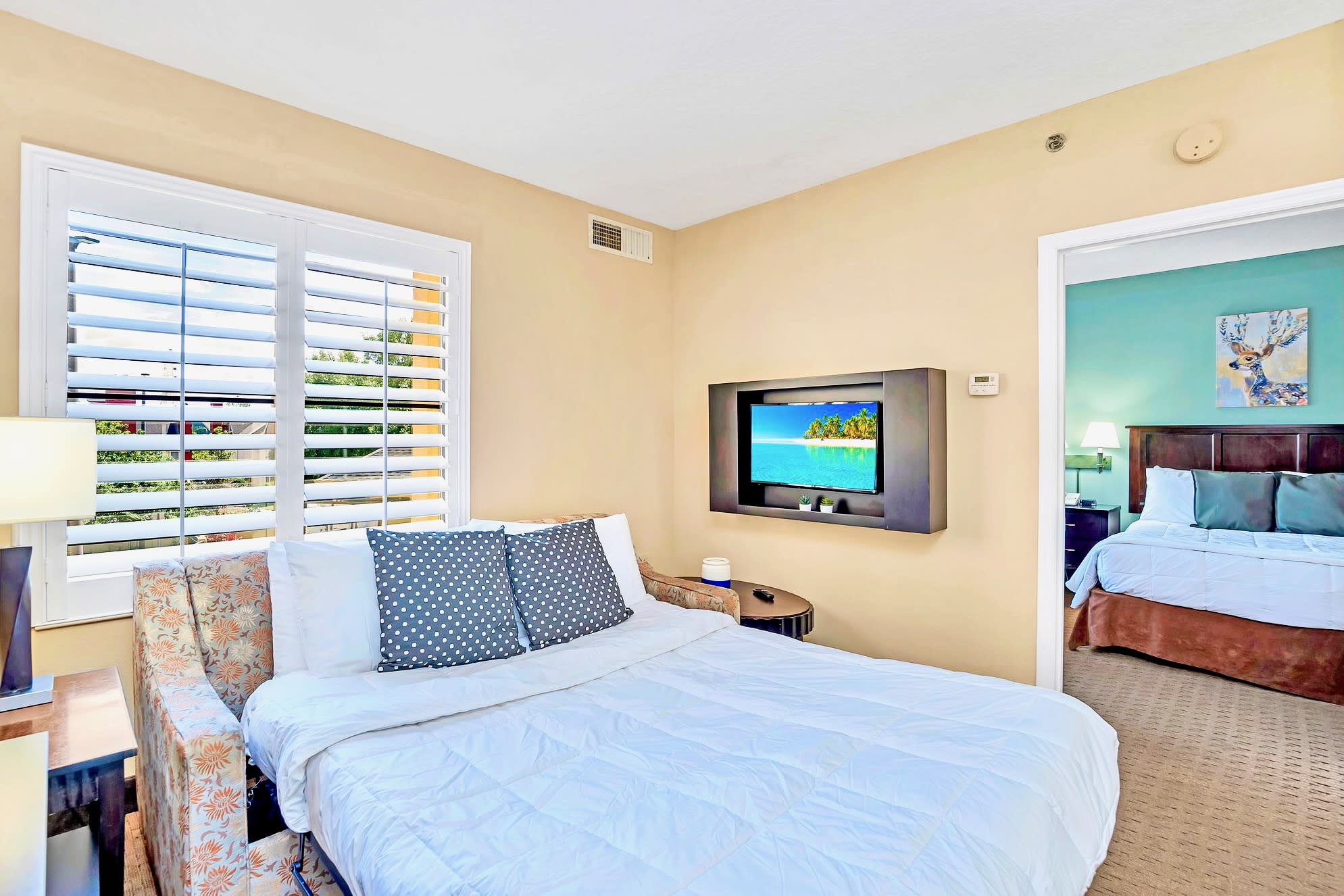A closer view of the living area, highlighting a pull-out sofa bed with patterned pillows and a wall-mounted TV displaying a beach scene. A window with white plantation shutters provides natural light, and an open doorway leads to an adjacent bedroom.