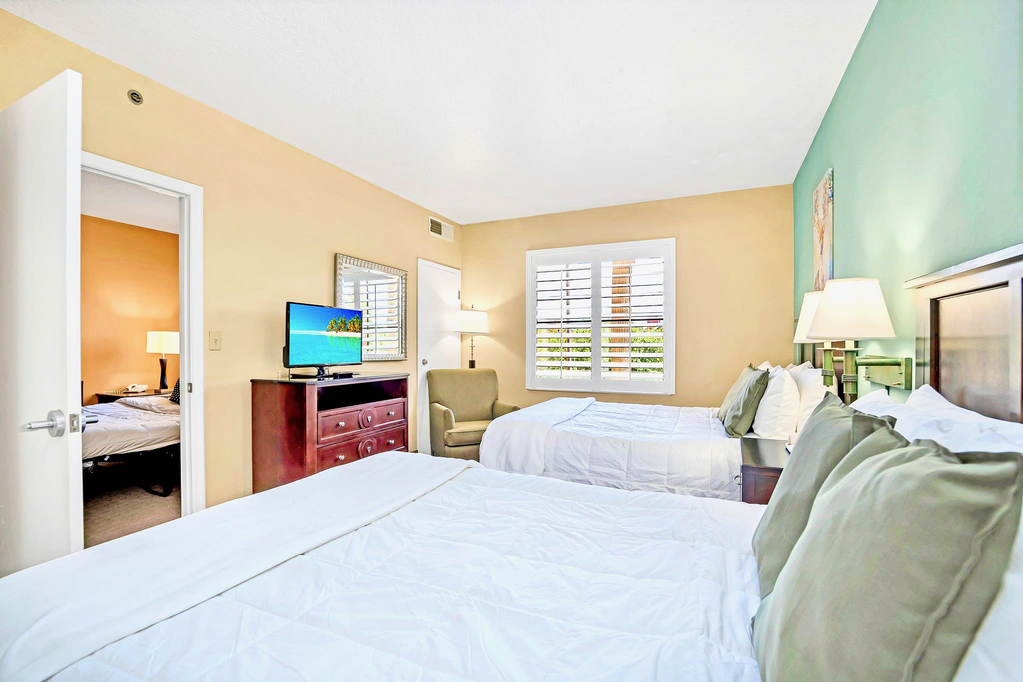 A bedroom featuring two beds with white bedding and green accent pillows, a wooden dresser with a TV, and a light-colored armchair. A window with white plantation shutters and a mirror above the dresser complete the space.