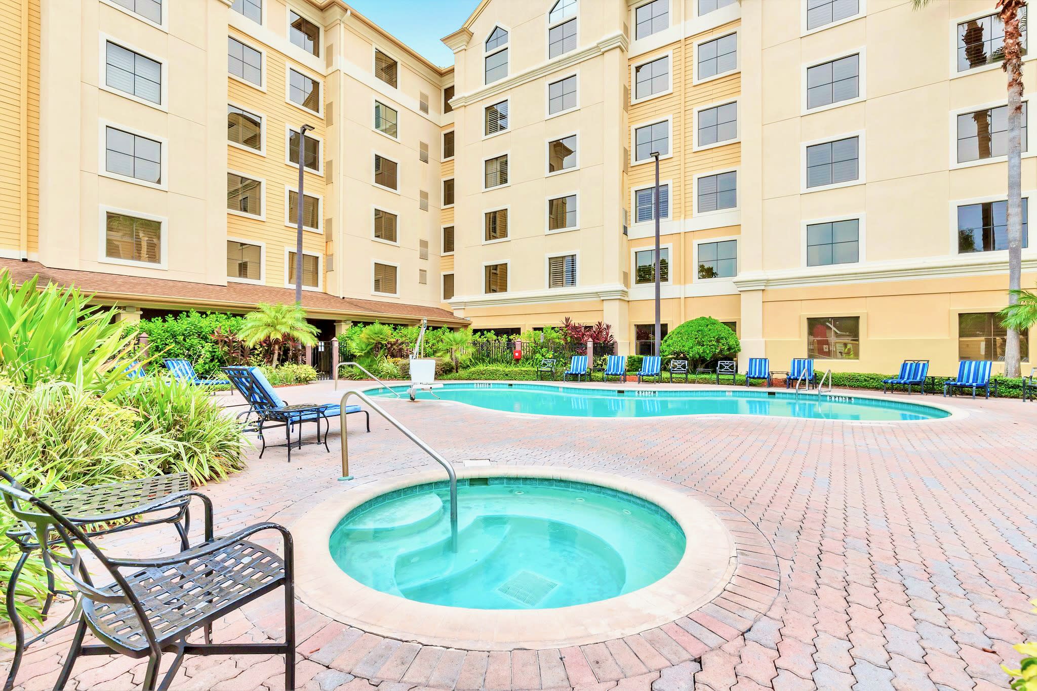 Outdoor swimming pool and circular hot tub set on a brick patio, surrounded by lounge chairs and lush green landscaping, with a multi-story hotel-style building in the background.