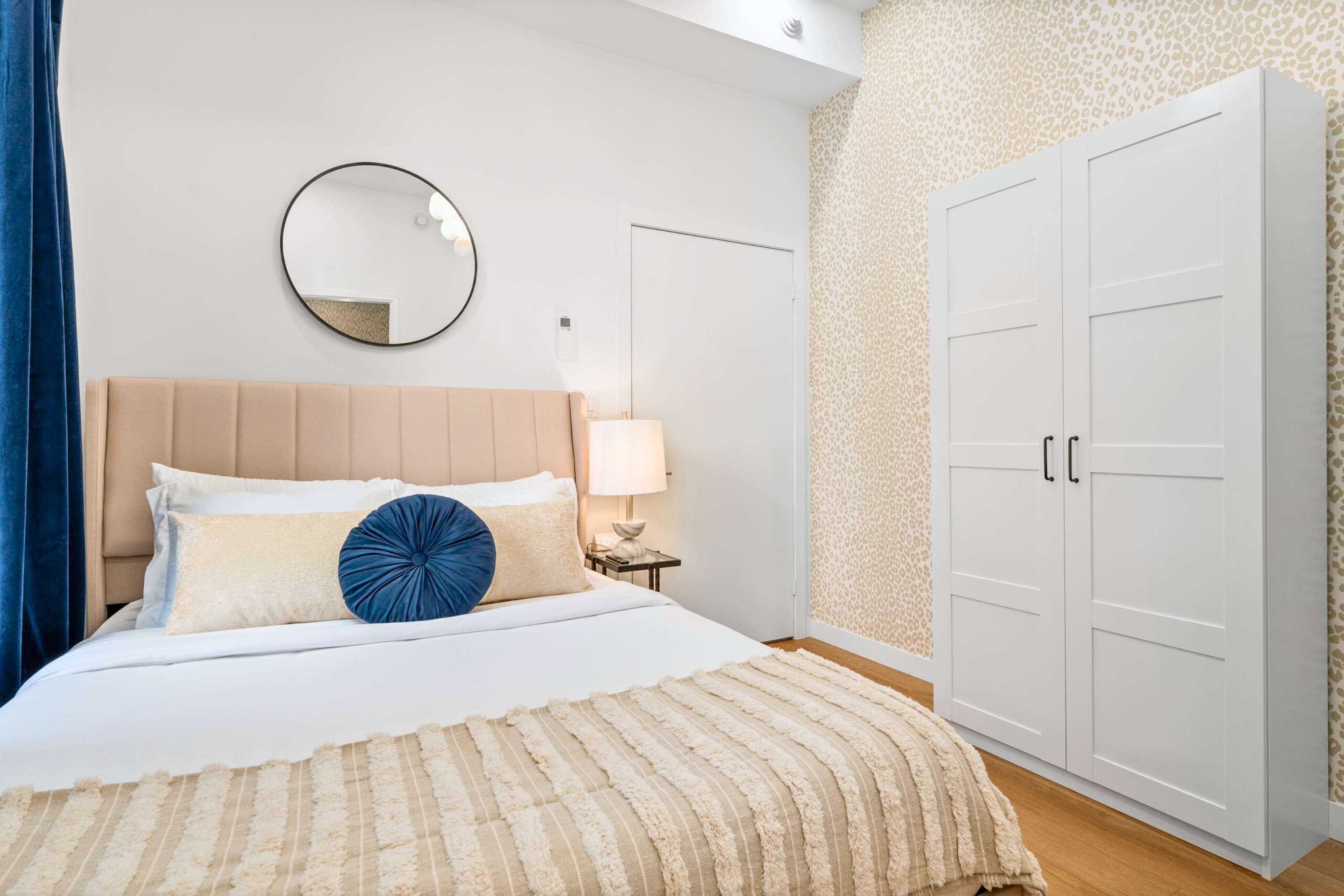 Cozy bedroom featuring a beige upholstered bed with blue and cream pillows, light wood flooring, a white wardrobe, and subtle patterned wallpaper on one accent wall.