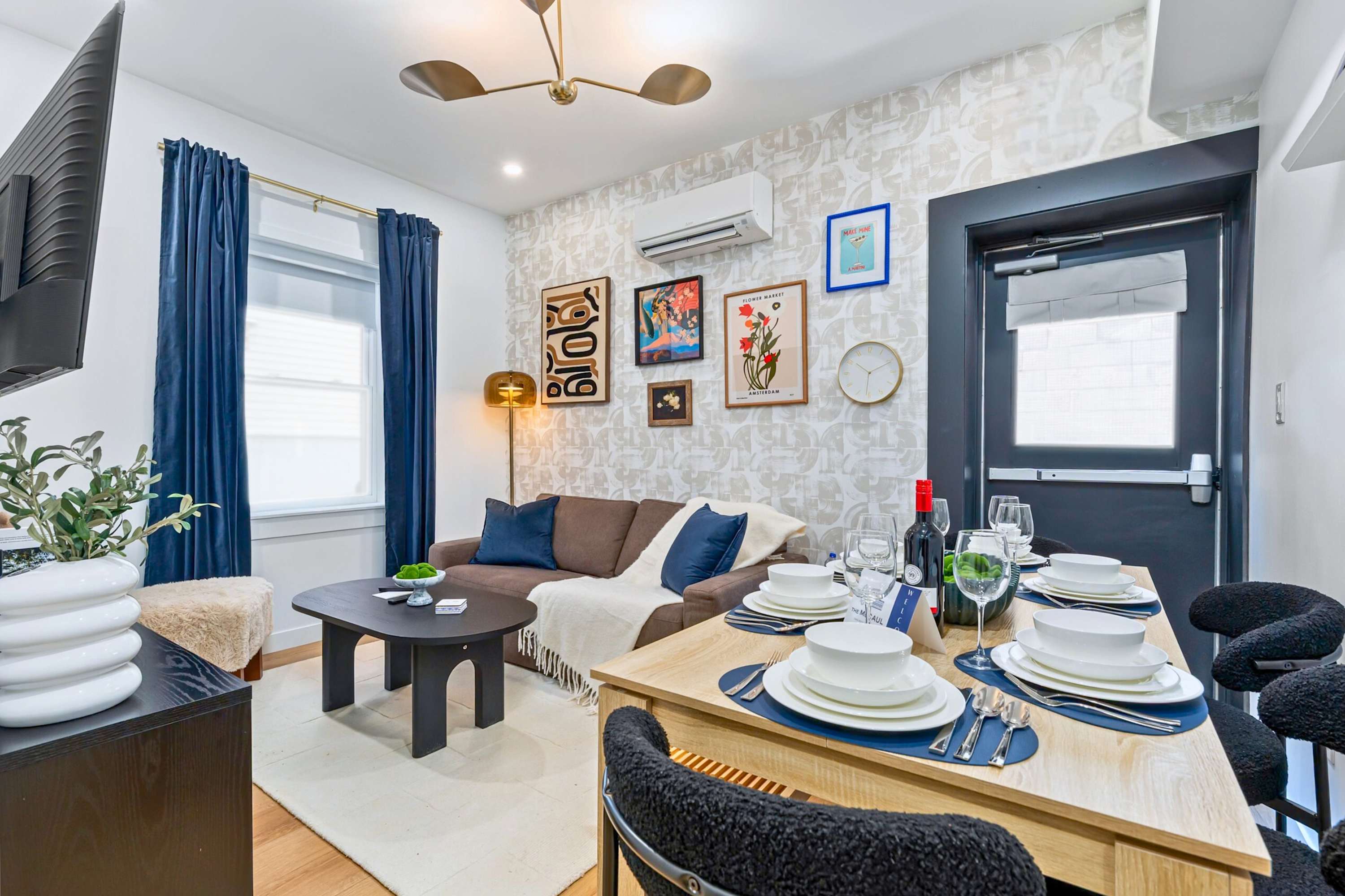 Open-concept living and dining area with a brown sofa, black coffee table, and a light wood dining table. The wall features patterned wallpaper and a gallery of framed art, with blue curtains by the window.
