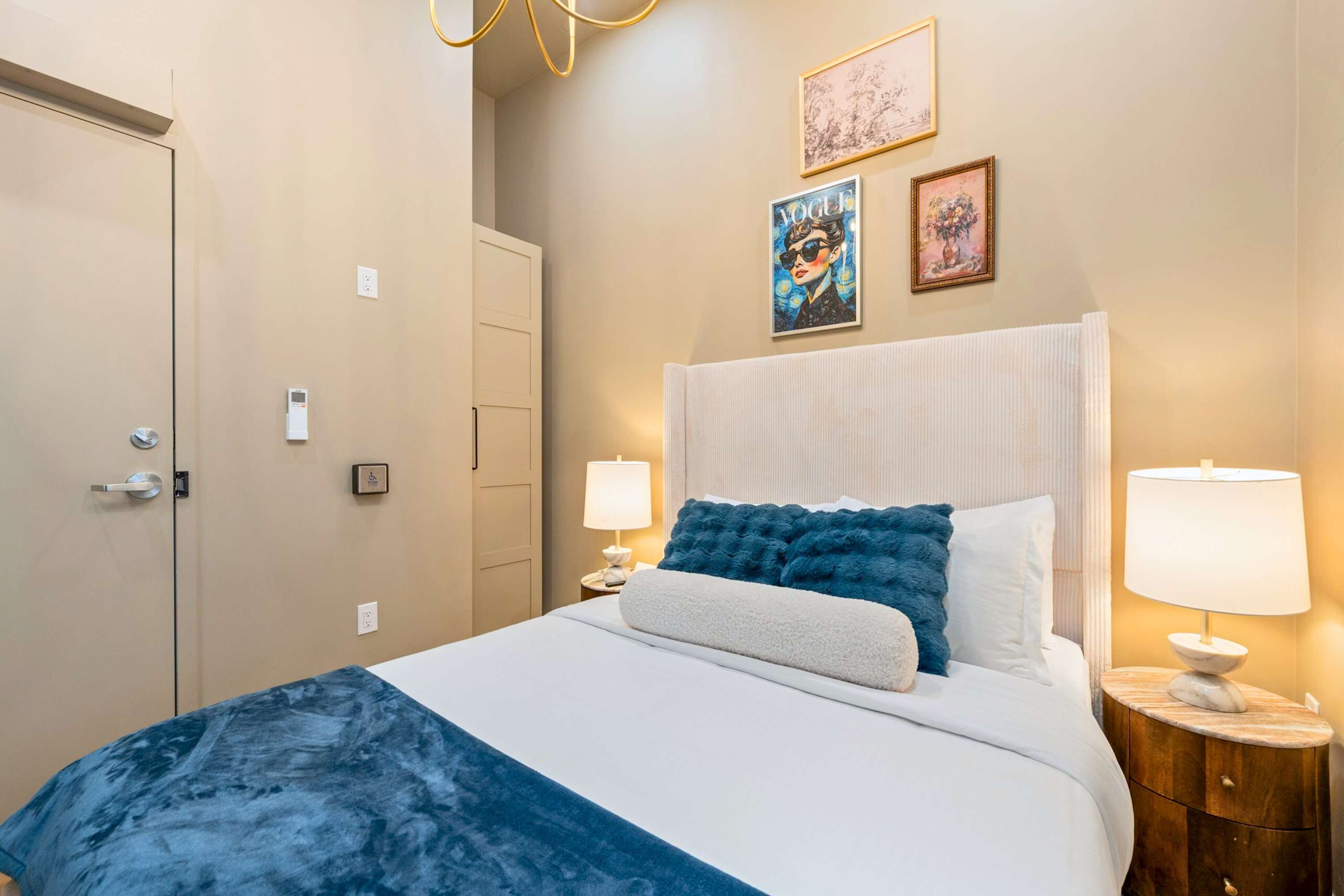 A cozy bedroom featuring a queen-sized bed with white linens and blue accents, flanked by a wooden nightstand with a lamp, and decorated with framed art and a Vogue poster on the beige wall.