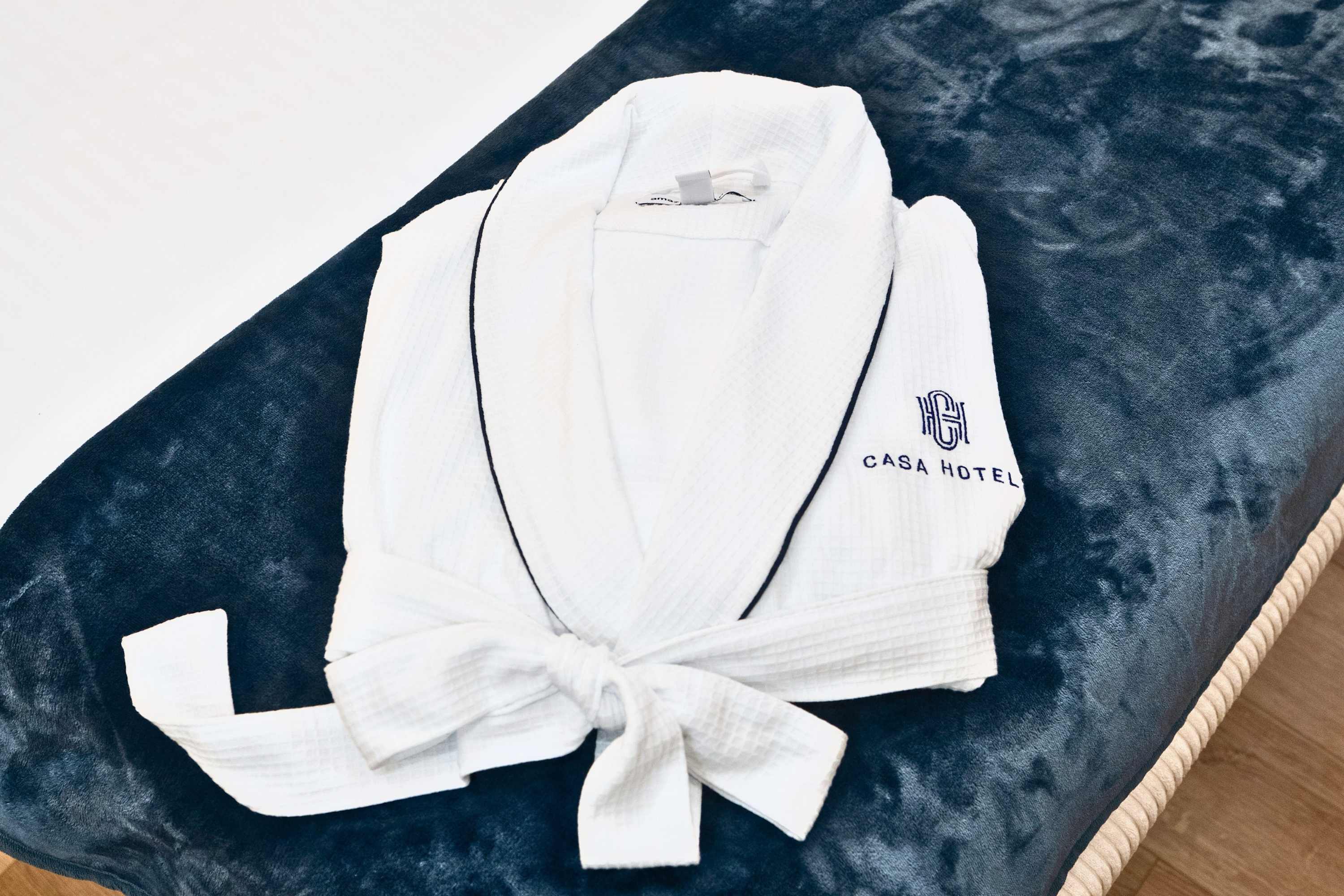 A white waffle-knit bathrobe with a dark blue trim and "CASA HOTEL" logo embroidered on the chest, neatly folded and placed on a plush blue blanket.