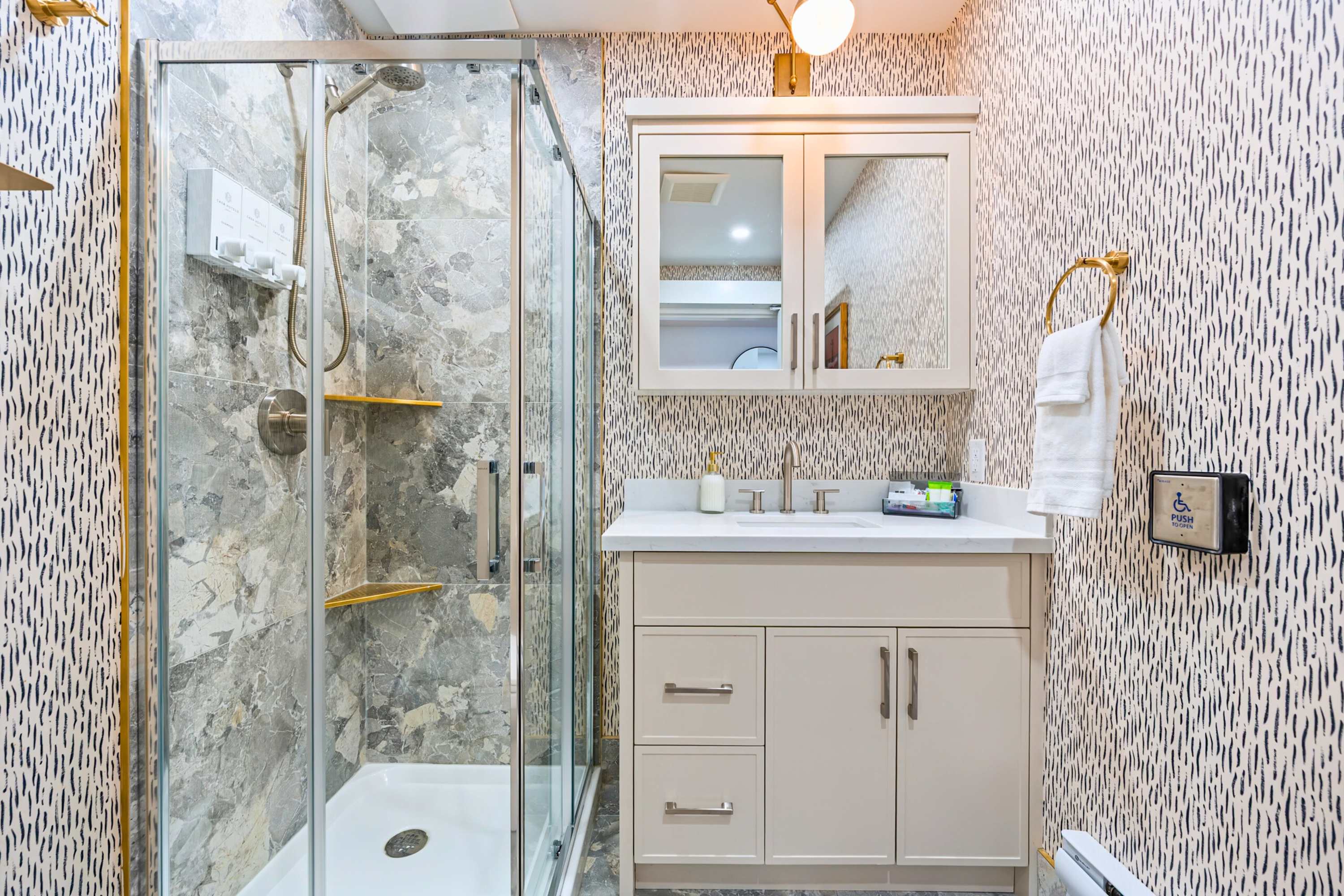 A modern bathroom featuring a glass-enclosed shower with grey marble-patterned tiles and gold accents, alongside a light grey vanity with a white countertop, sink, and mirrored medicine cabinet.