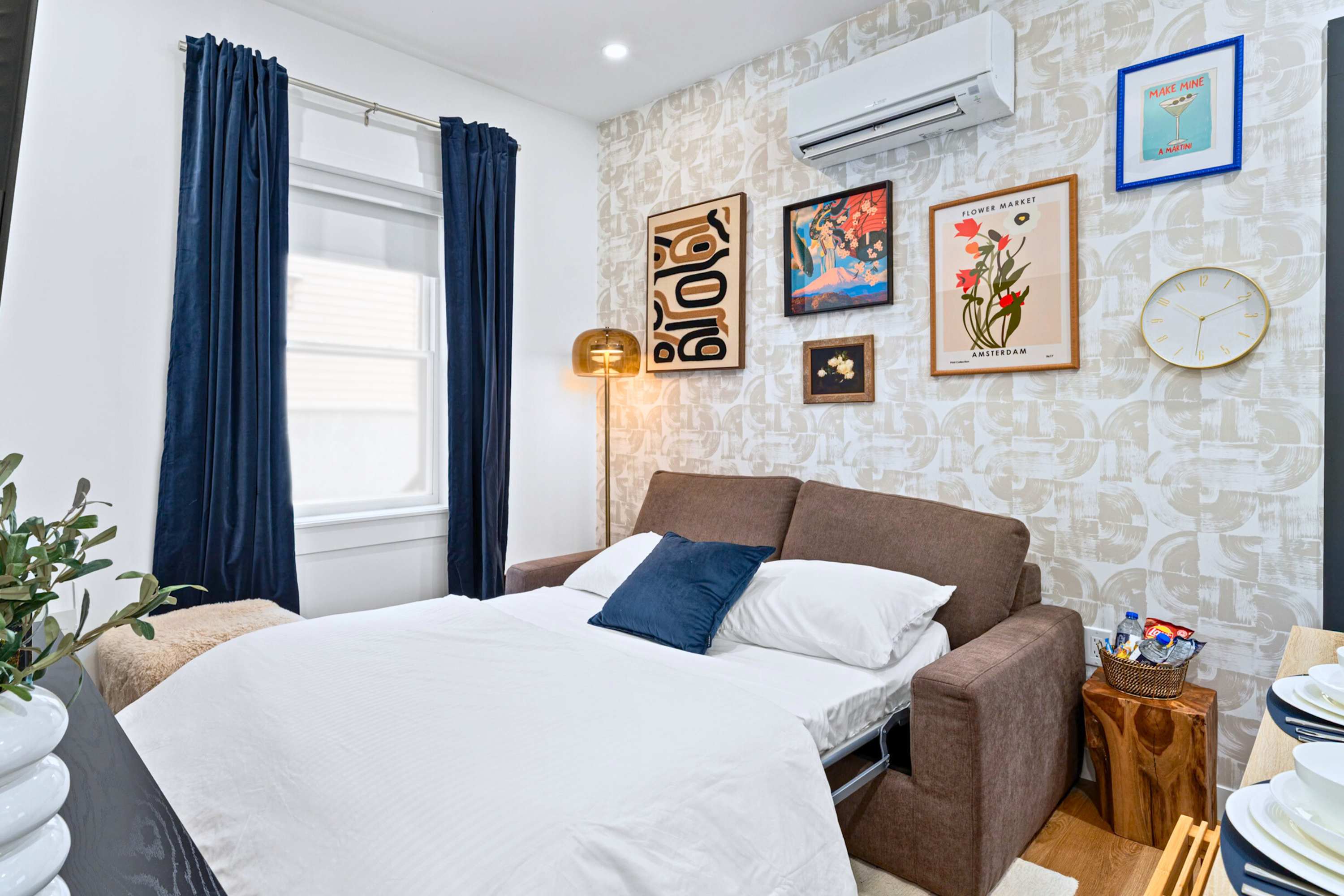 Living room transformed into a sleeping area with the brown sofa converted into a pull-out bed, dressed with white sheets and a blue pillow. A black coffee table and patterned wallpaper with artwork are visible.