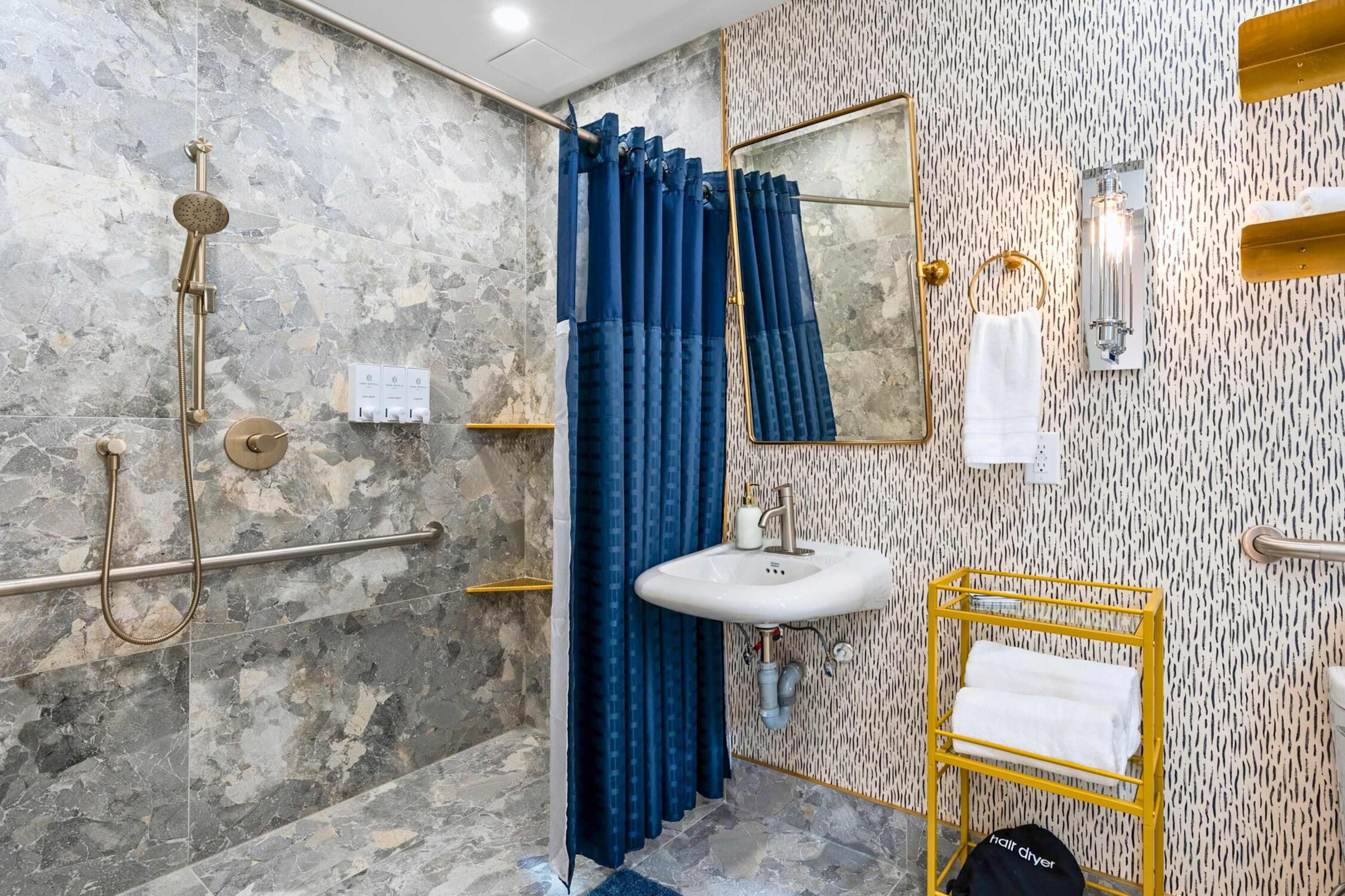 Modern bathroom with gray marble-patterned tiles on the floor and shower walls, a walk-in shower with a blue curtain, a small wall-mounted sink, and patterned wallpaper.