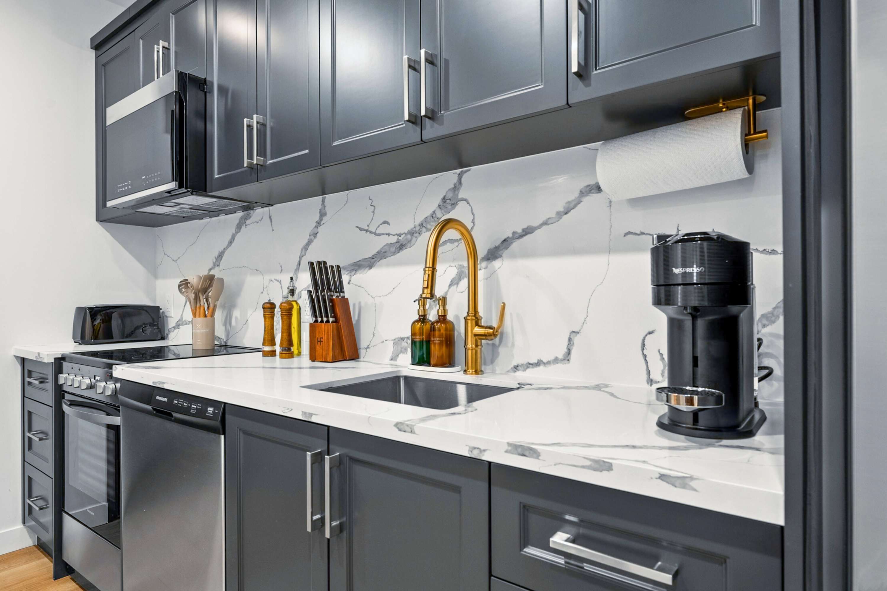 Detailed view of the kitchen sink area featuring a gold-toned faucet, white quartz countertops, a white marble-patterned backsplash, and a black Nespresso machine.