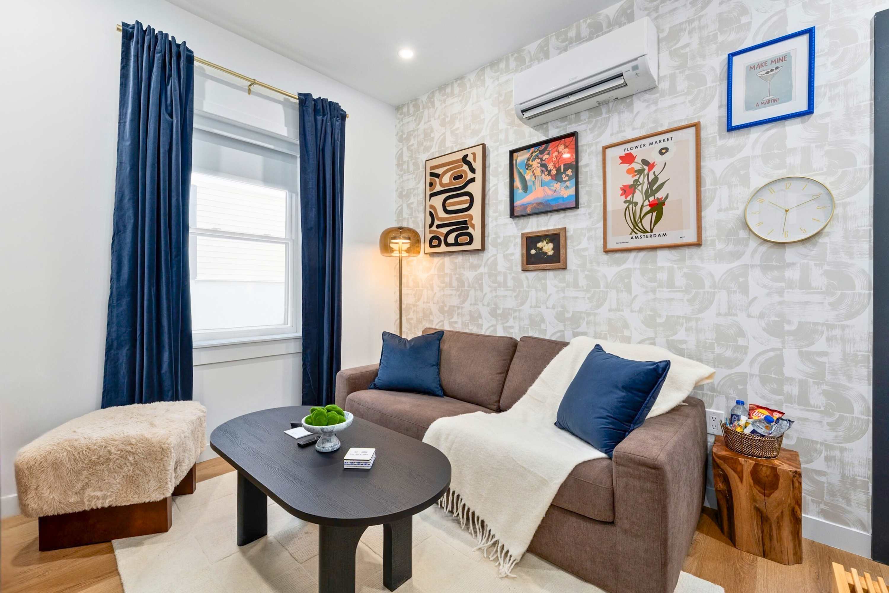 Inviting living room with a brown sofa, plush throw blanket, and blue accent pillows. A black oval coffee table sits on a light area rug, and a gallery wall with diverse artwork decorates the patterned wallpaper.