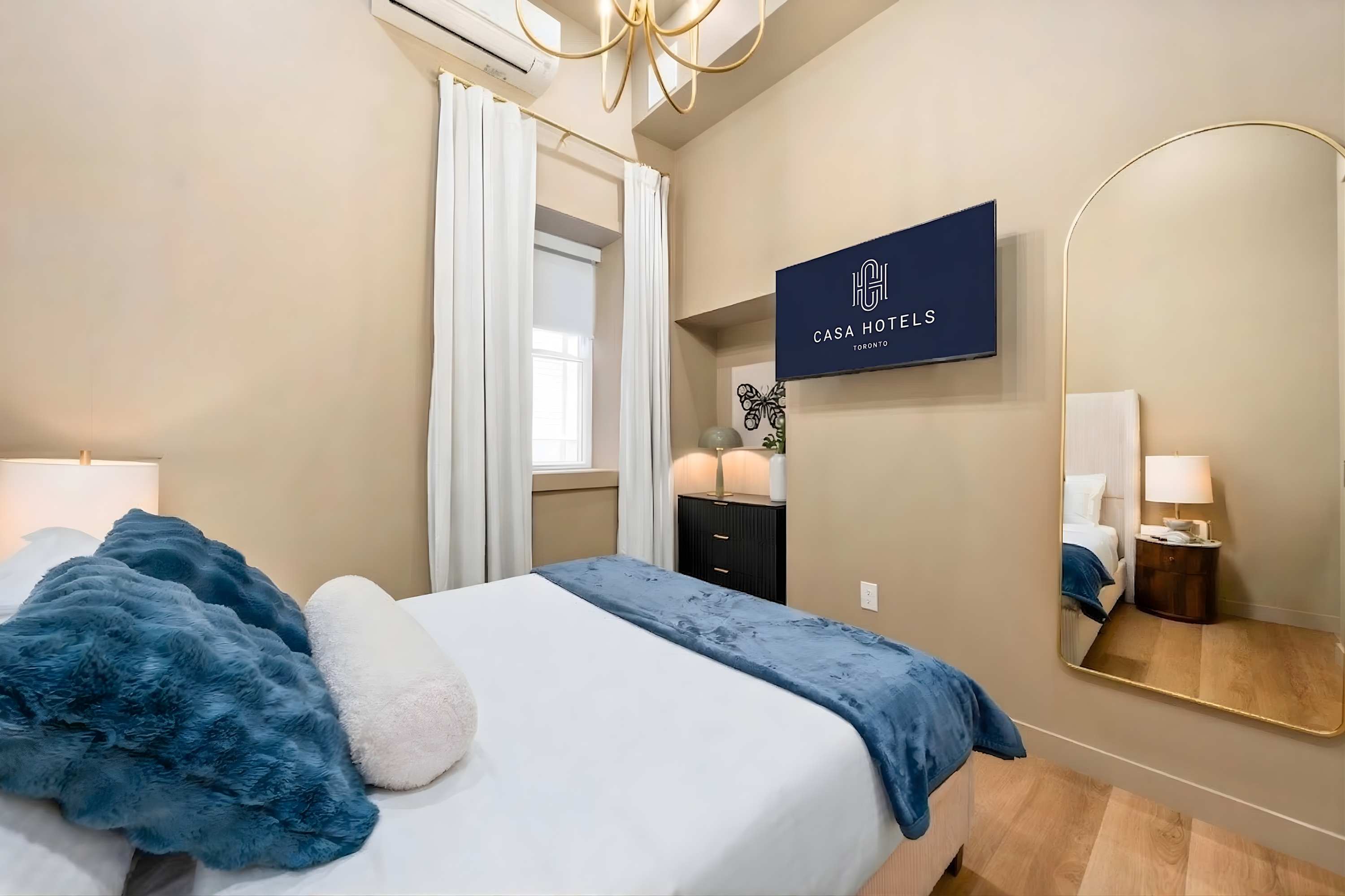 A different angle of the bedroom, showcasing a wall-mounted TV with "CASA HOTELS TORONTO" logo, a full-length arched mirror, and a window with white curtains, all contributing to a modern and bright aesthetic.