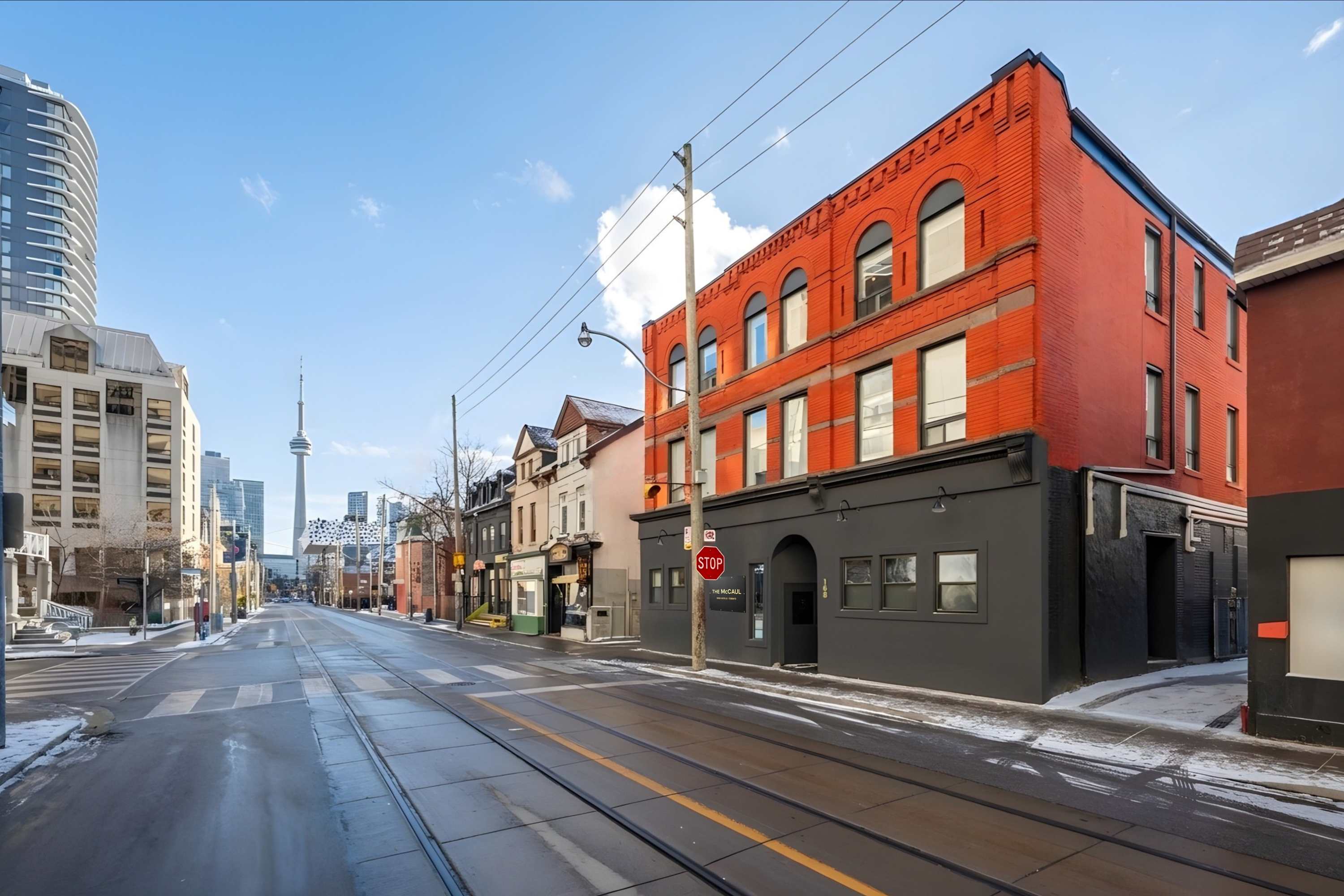 A street-level view of a city street with a red brick building on the right, featuring a dark grey lower facade with "THE MCCAUL" sign, and the CN Tower visible in the distant skyline under a clear sky.