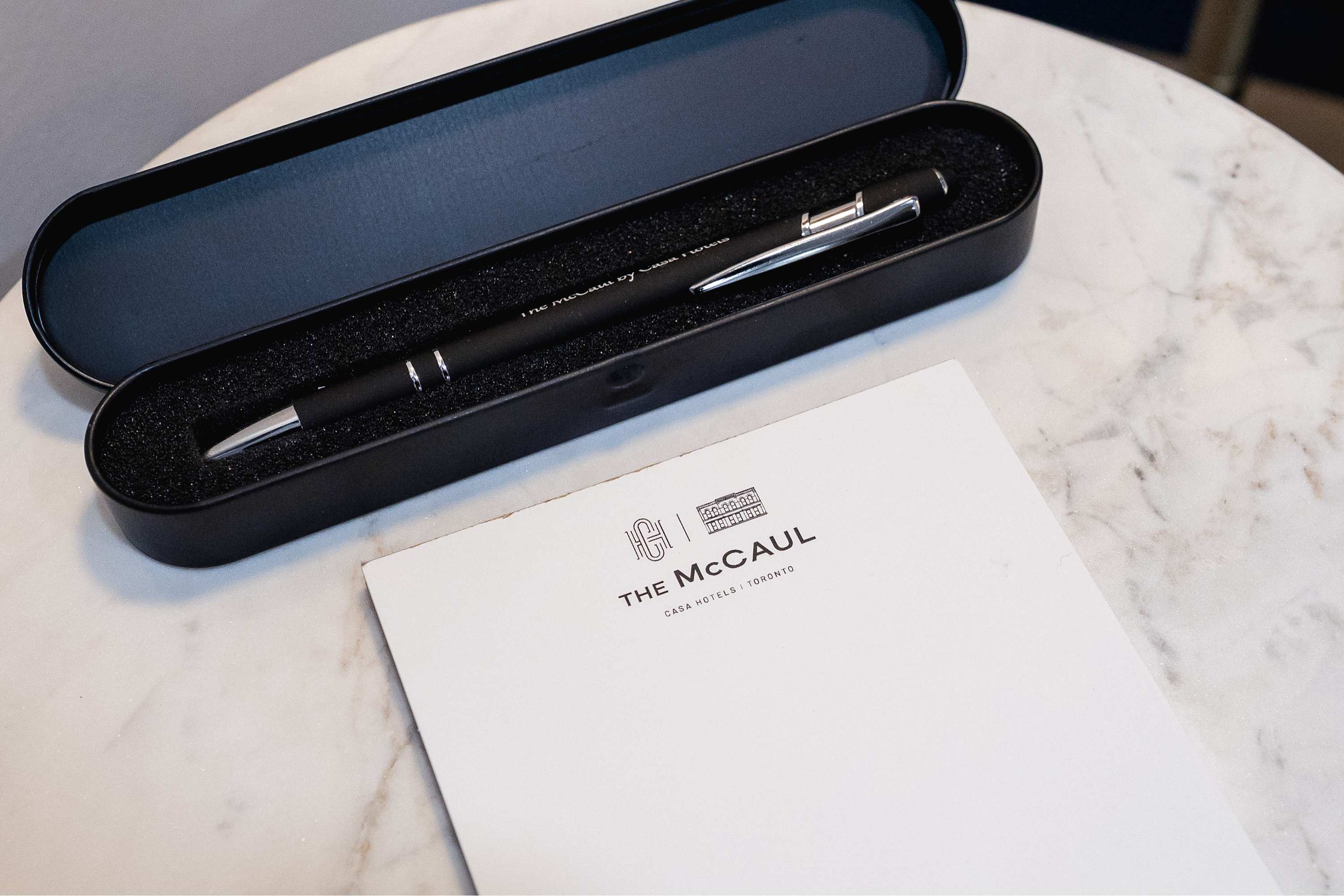 Close-up of a black pen in a black case next to a white notepad featuring "THE MCCAUL CASA HOTELS | TORONTO" logo, resting on a light marble-patterned surface.
