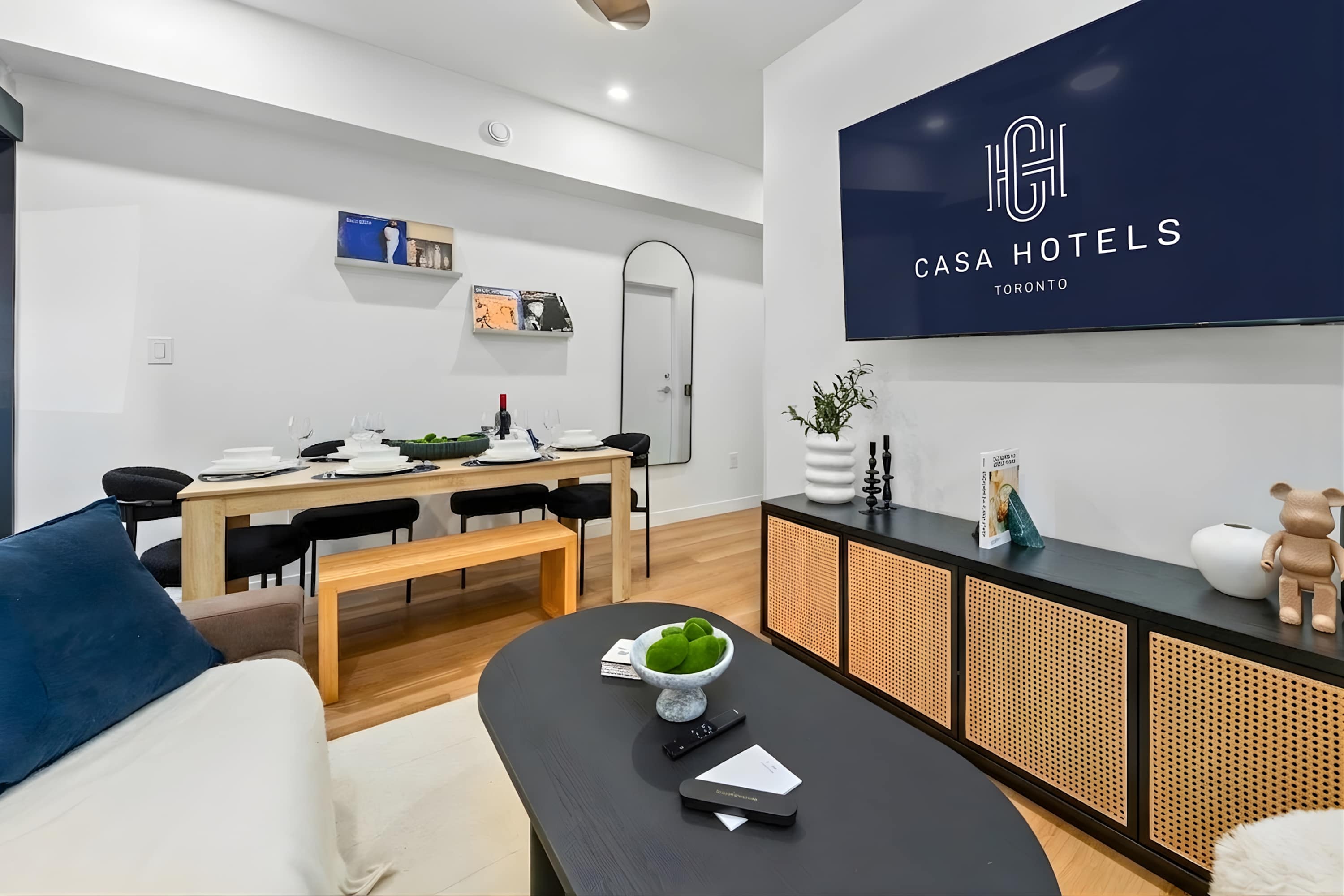 Open-concept living and dining area featuring a brown sofa, black coffee table, and a light wood dining table with black chairs. A large wall-mounted TV is visible, and decorative records are displayed on wall shelves.