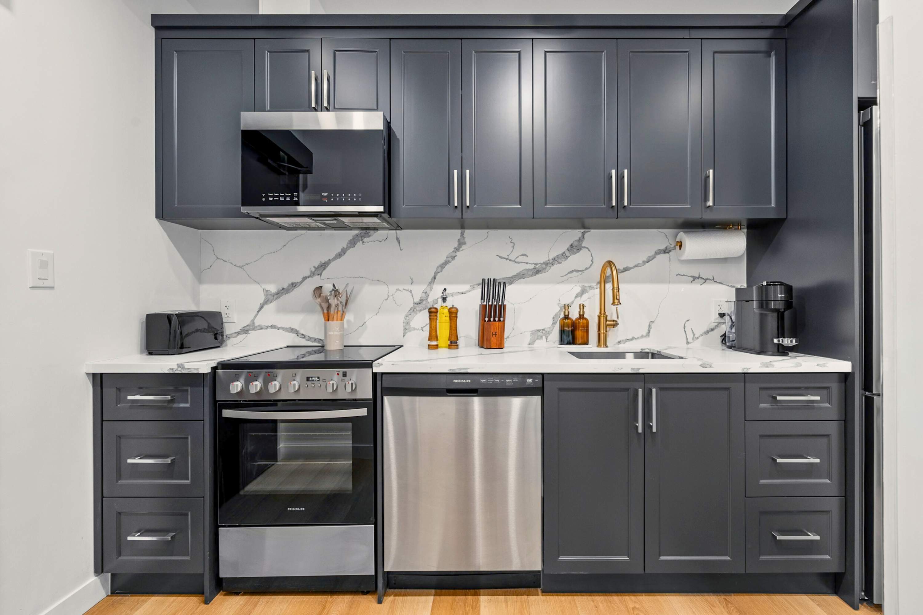 Compact and modern kitchen with dark gray shaker cabinets, white quartz countertops, a white marble-patterned backsplash, and stainless steel appliances including an oven, dishwasher, and microwave.