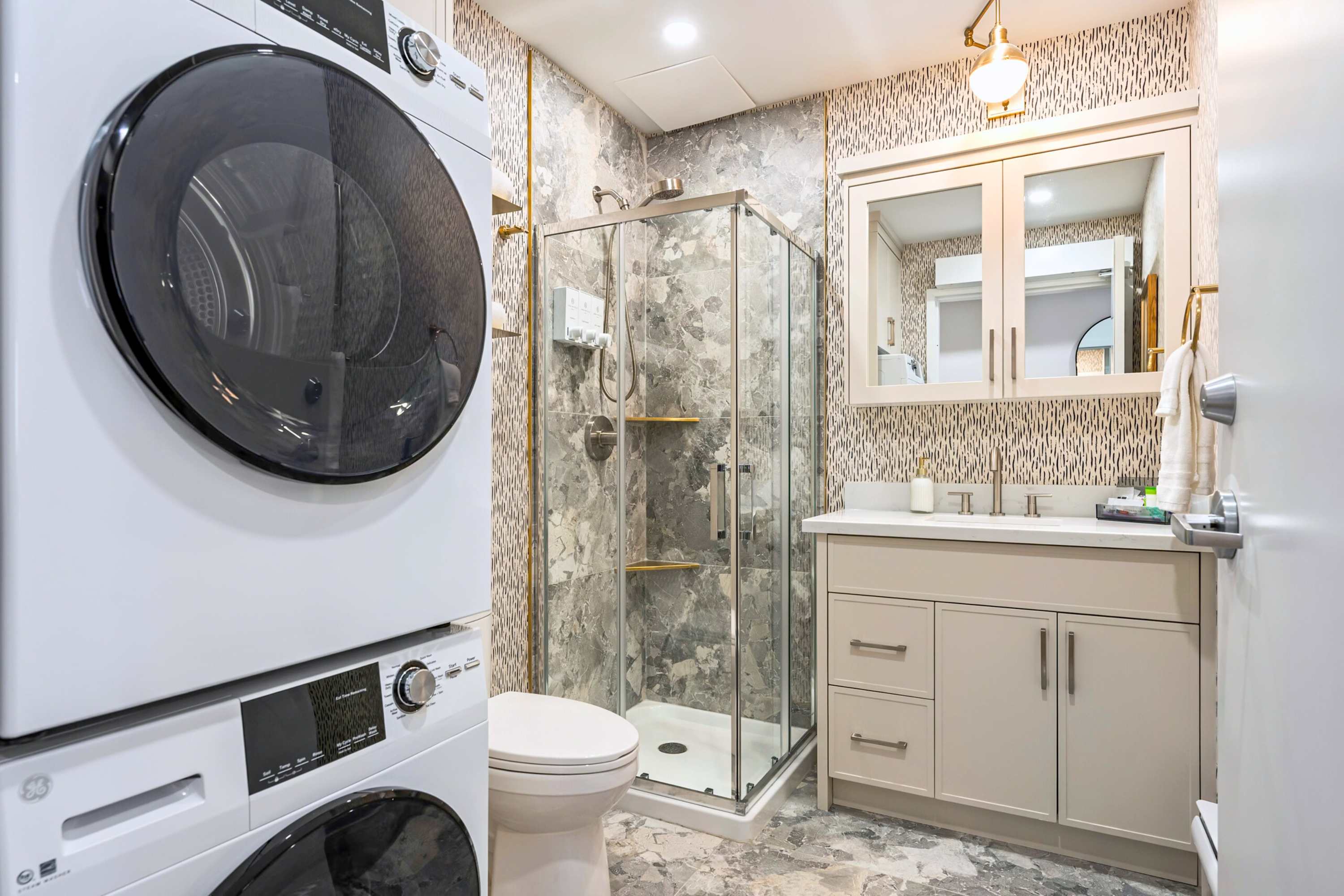 A wider view of the bathroom, revealing a stacked washer and dryer unit next to the toilet, complementing the glass-enclosed shower and vanity, all set against patterned wallpaper and grey tiled flooring.